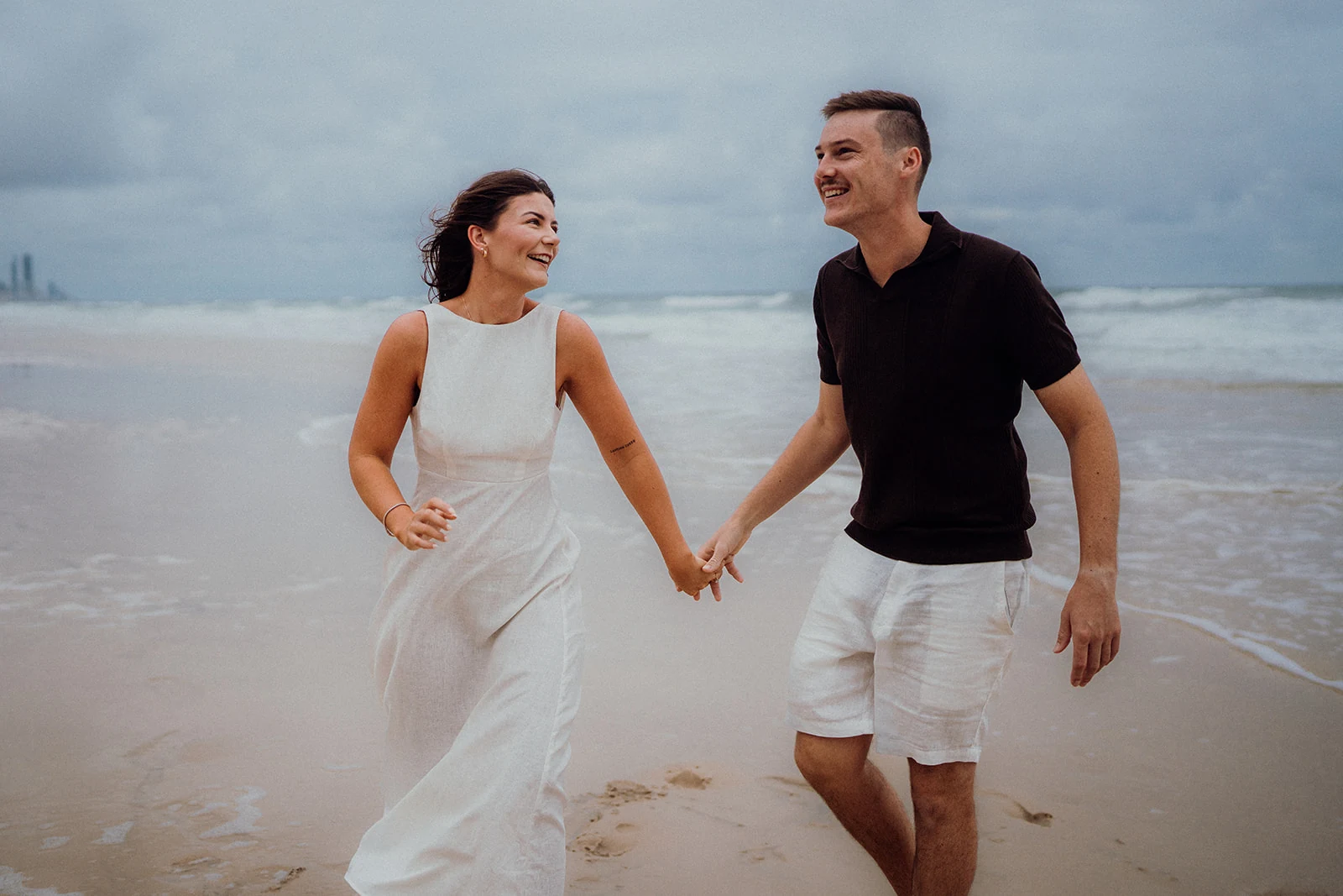 A couple laugh and hold hands while walking beside the waves on a beach.