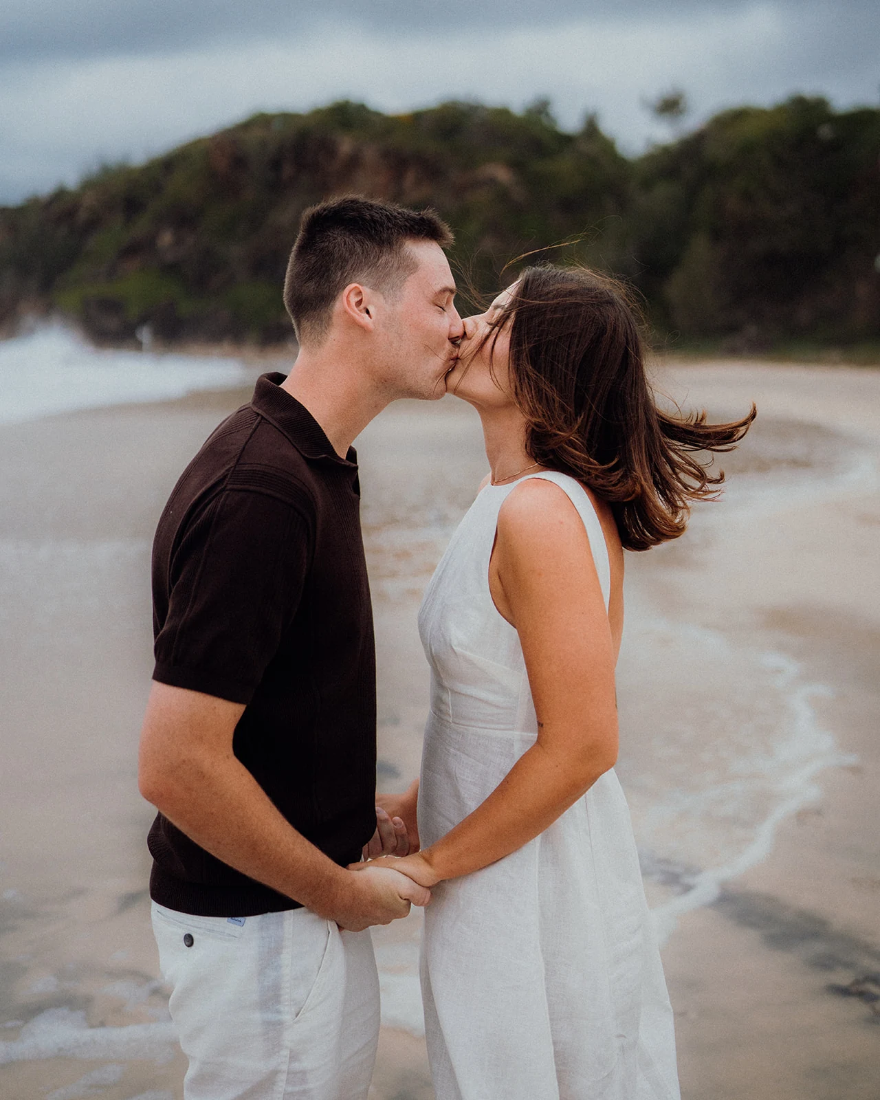 A couple hold hands and kiss beside the sea as the wind lifts her hair.