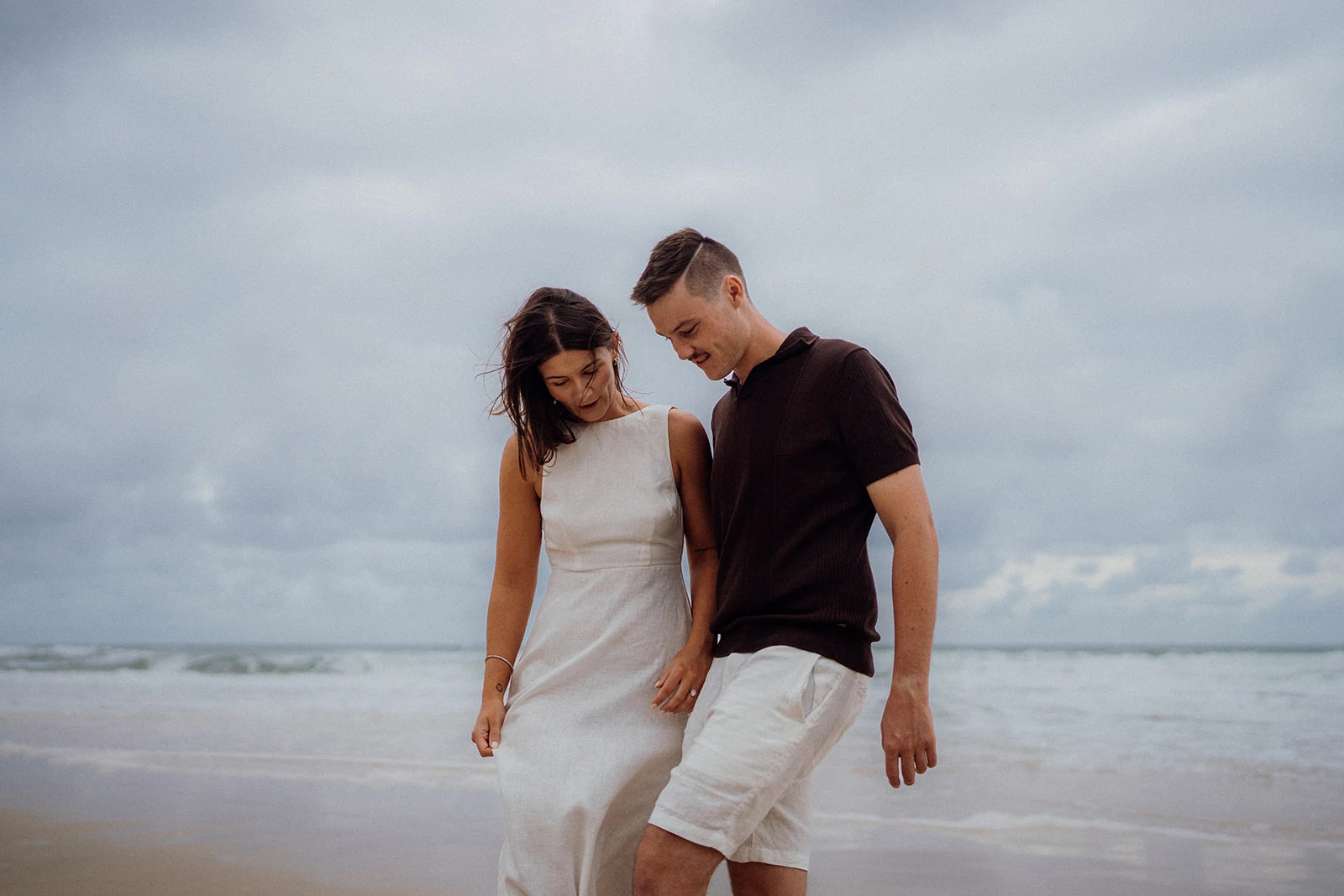 A couple walk close together through shallow water beneath a cloudy sky.