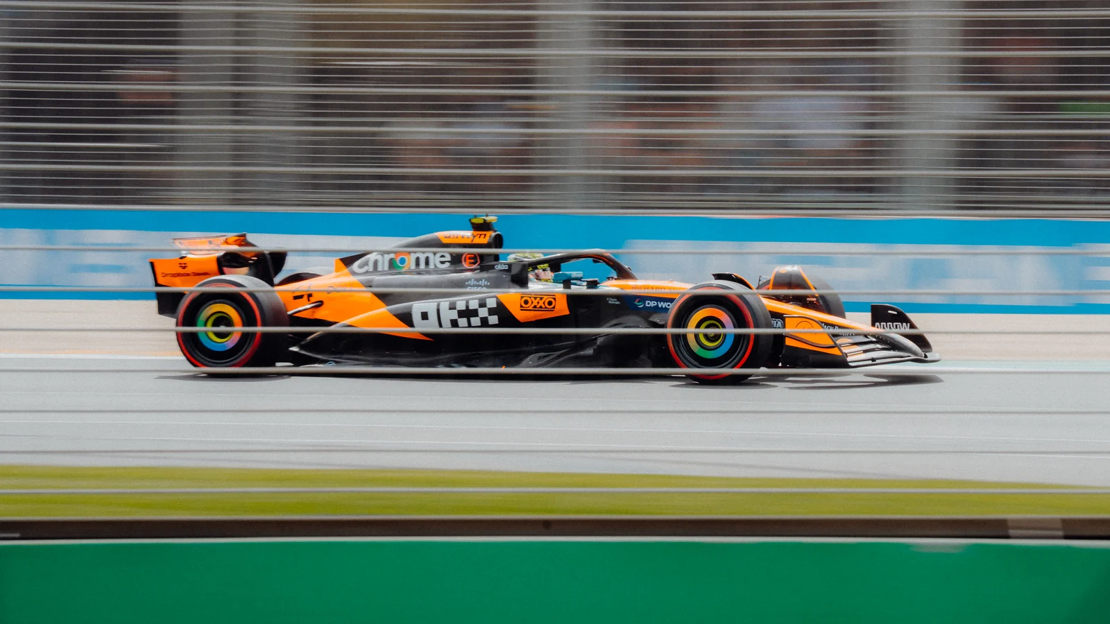 An orange Formula 1 car speeds past a blurred grandstand and trackside fence.