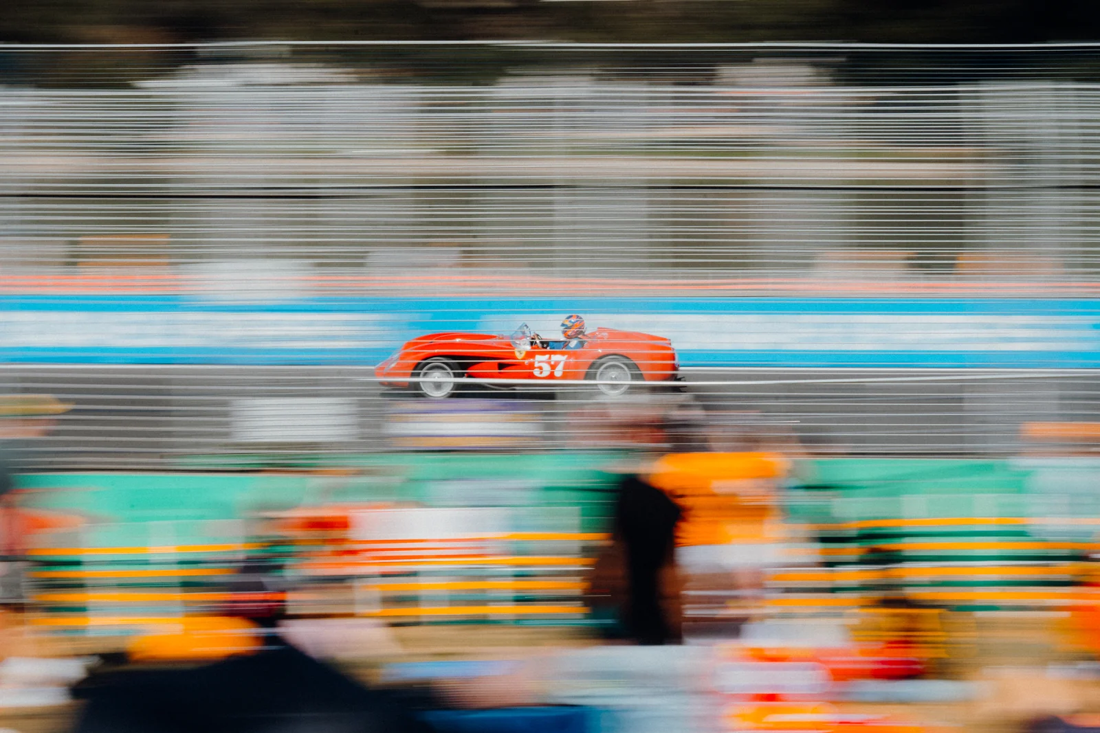 A red open-top historic racing car passes blurred spectators beside the circuit.