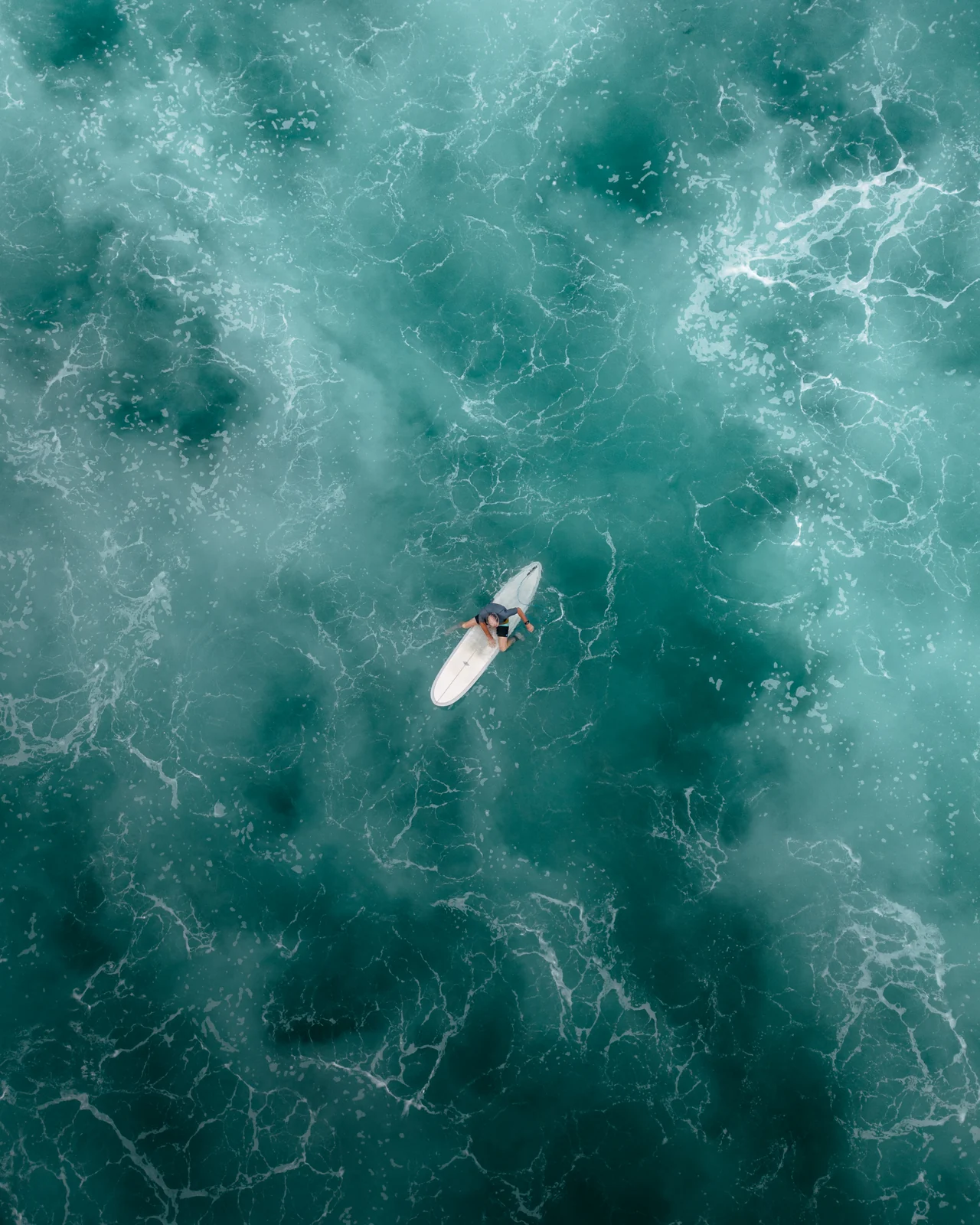 A surfer sits on a white board surrounded by turquoise water and foam, viewed directly from above.