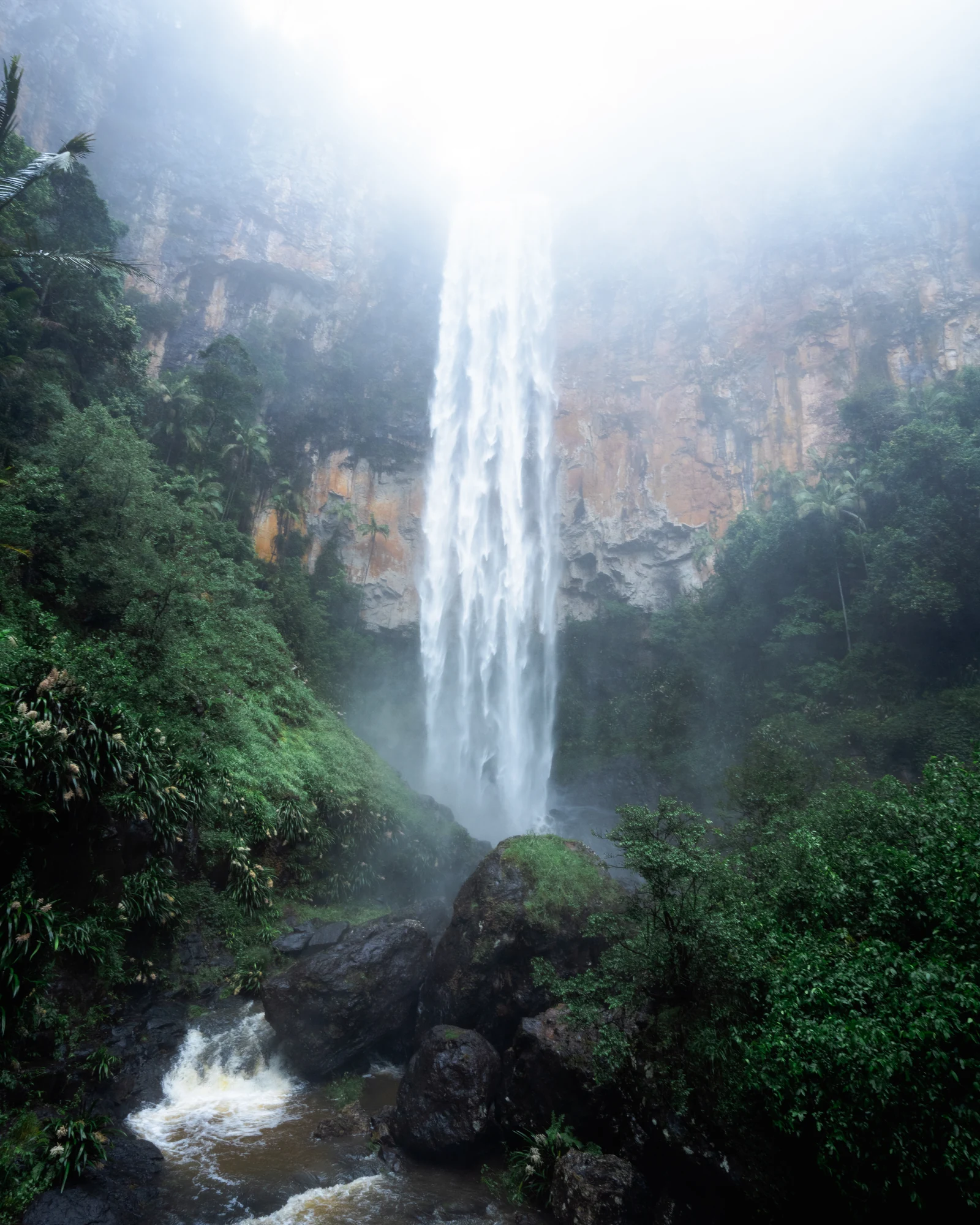 A tall waterfall drops through mist beside an orange cliff into a rocky forest stream.