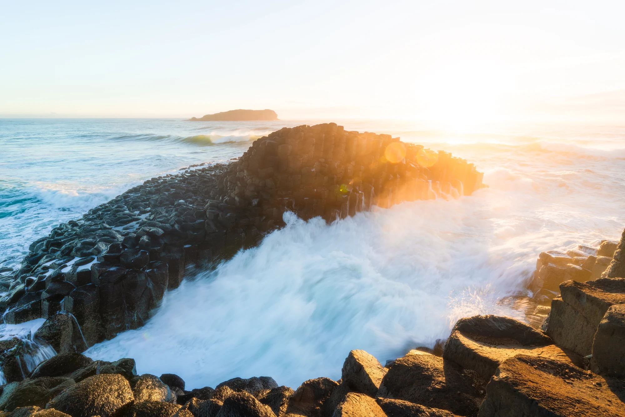 Sunrise lights angular coastal rocks as white water surges through a dark inlet.