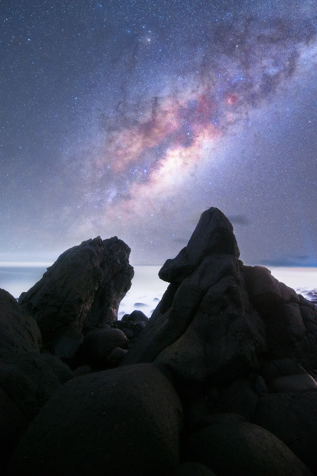 The Milky Way glows above jagged rocks on a dark coastline.