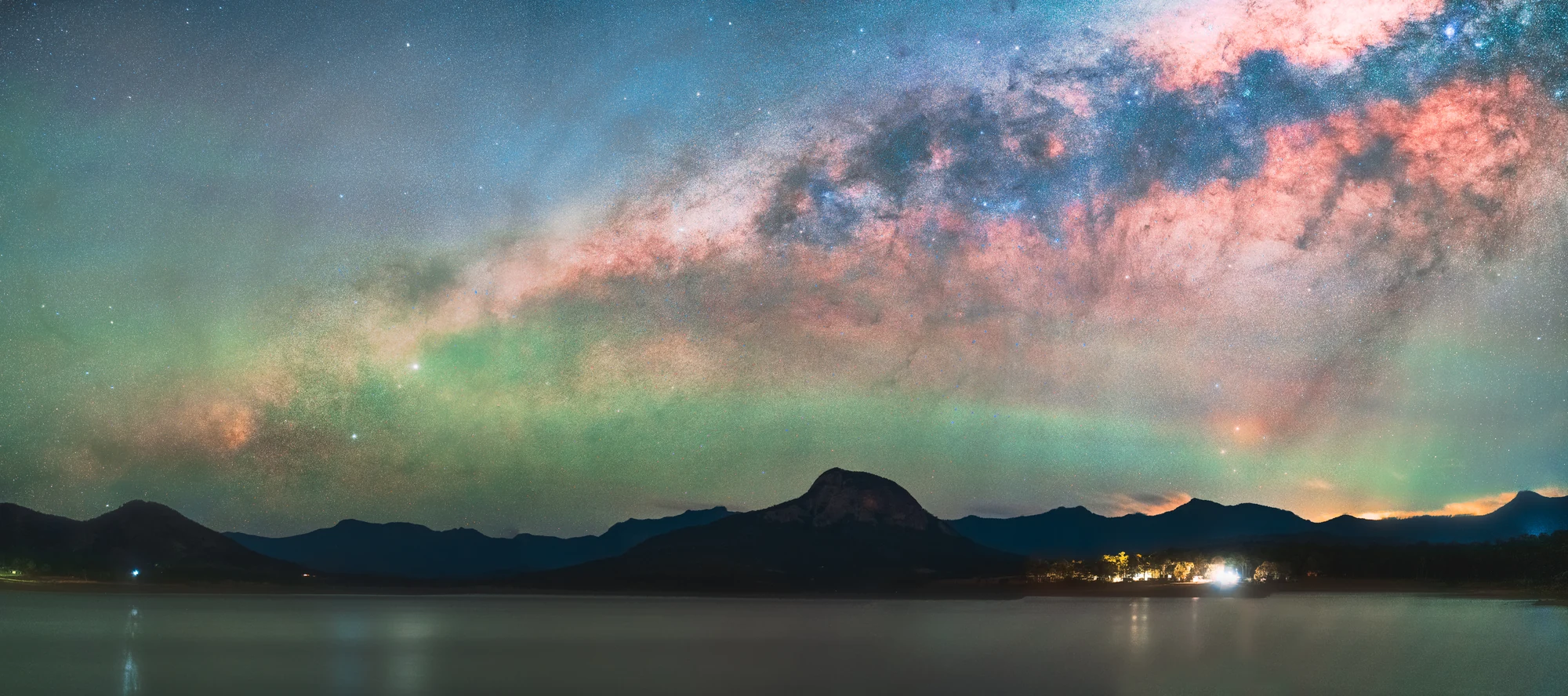 A luminous arc of the Milky Way spans a mountain lake beneath a pink and green night sky.