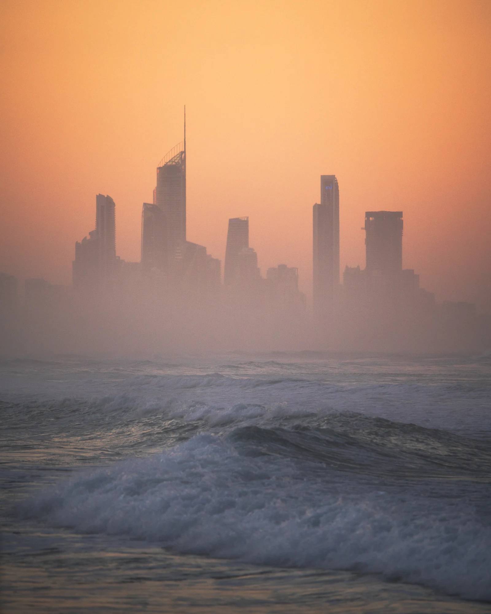 Ocean waves lead toward the Gold Coast skyline silhouetted in warm orange haze.