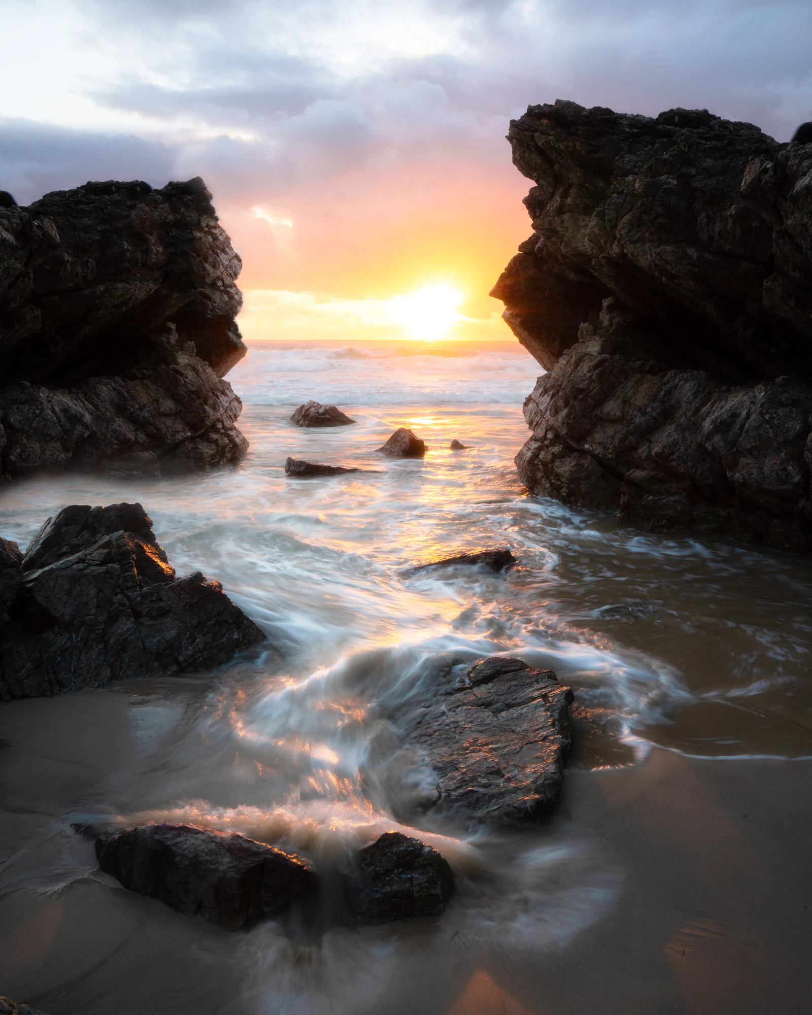 A low sun shines between jagged coastal rocks above trailing white water.