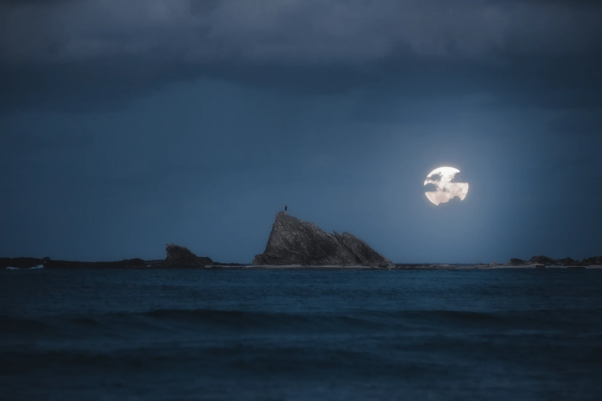 A pale moon hangs above jagged coastal rocks and a dark blue sea.