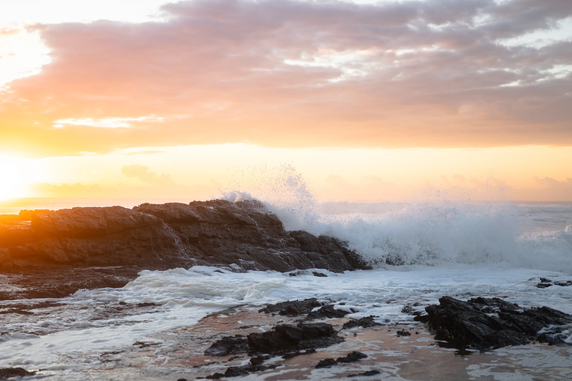 A wave breaks across coastal rocks beneath pink sunrise clouds.