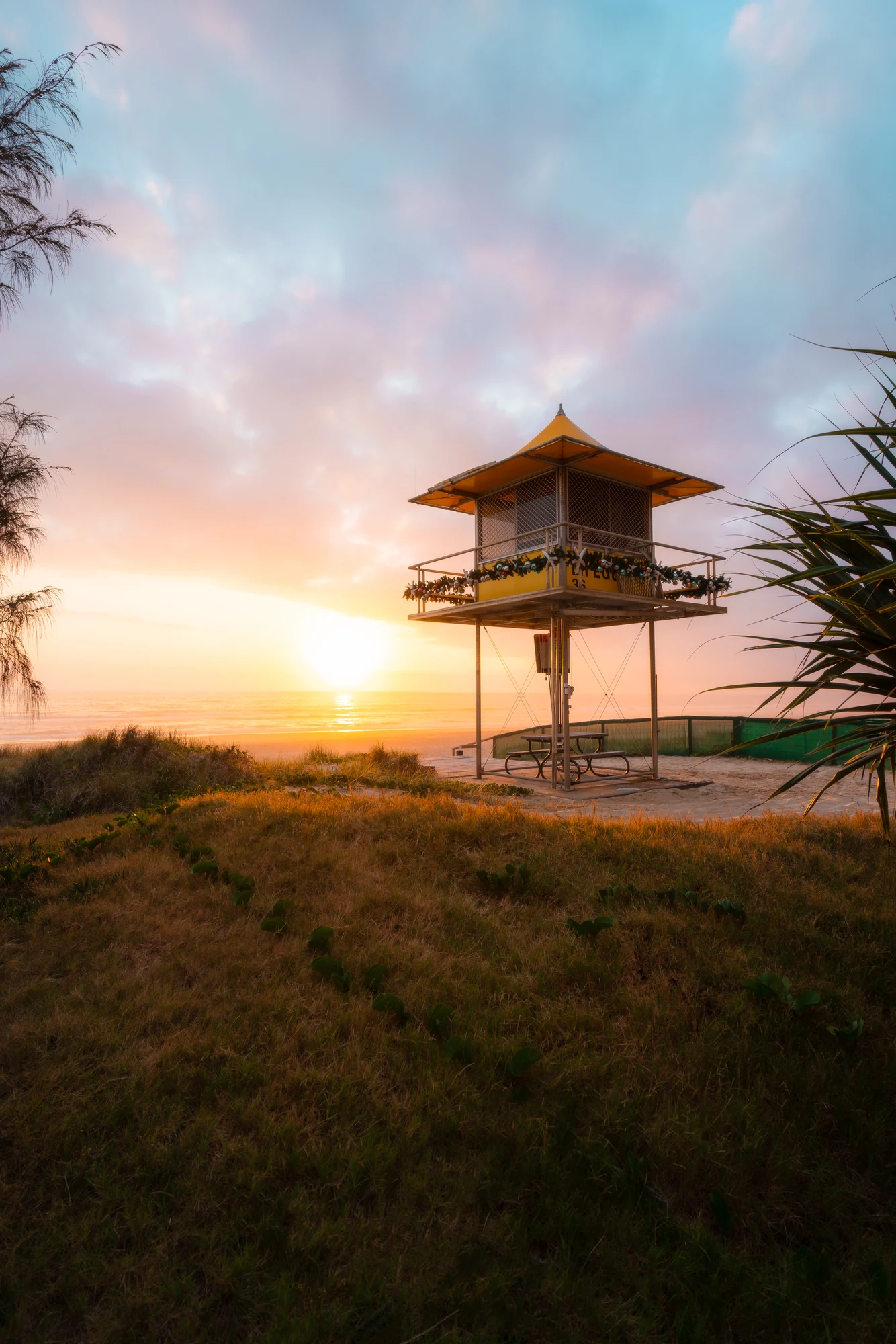 A yellow lifeguard tower stands above beach grass beneath pink and blue clouds.