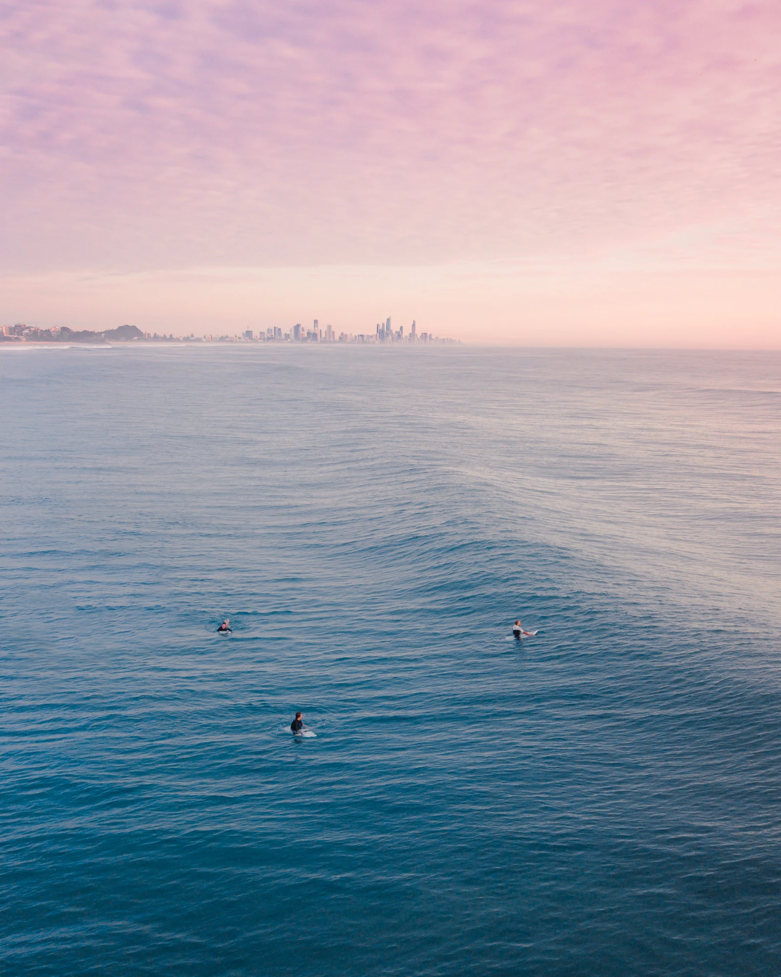 Three surfers wait in a wide blue sea beneath a pastel sky and distant city skyline.