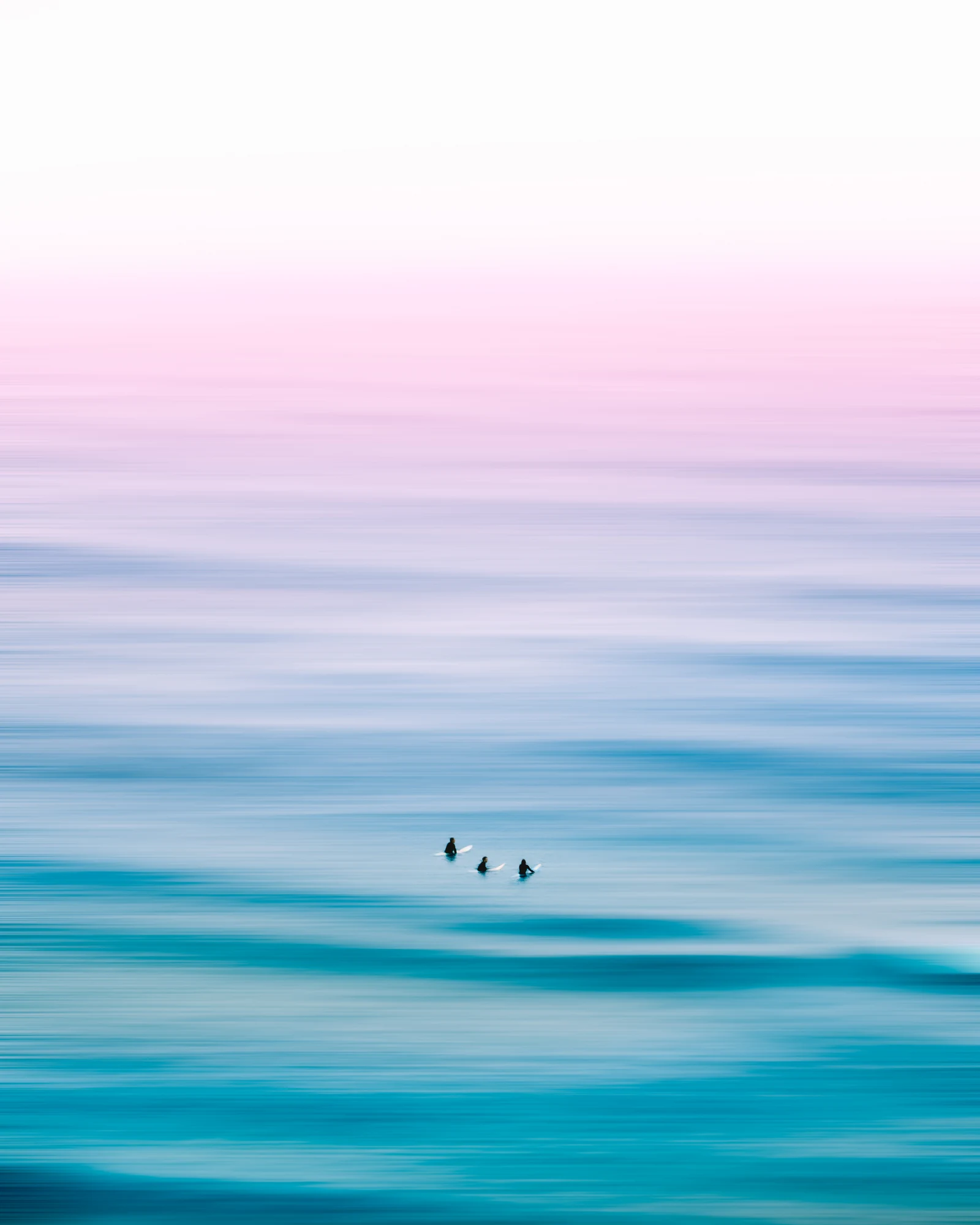 Three small surfers sit among soft blue and pink bands of ocean water.