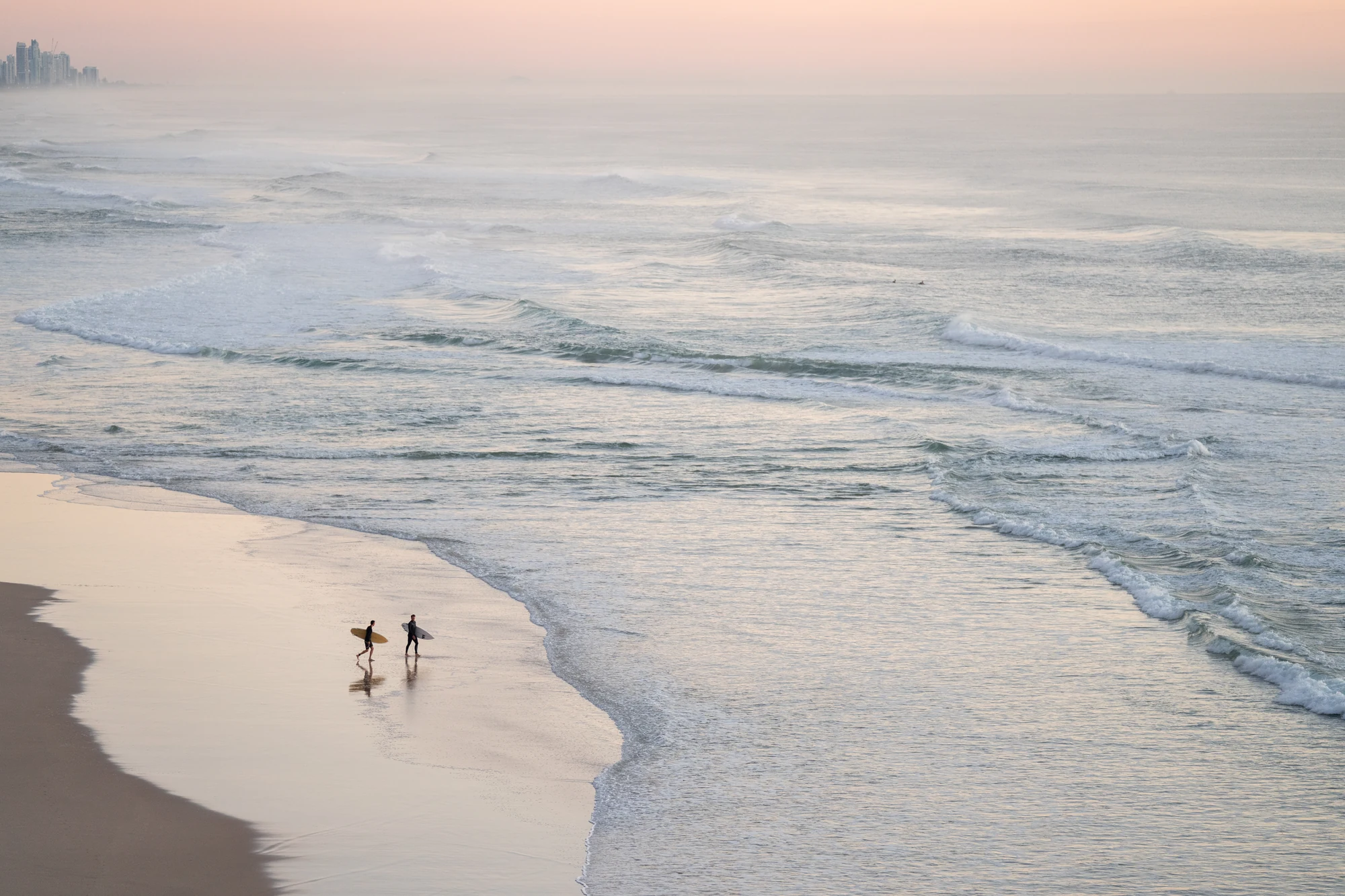 Two small surfers stand beside a curving shoreline reflected in pale evening sand.