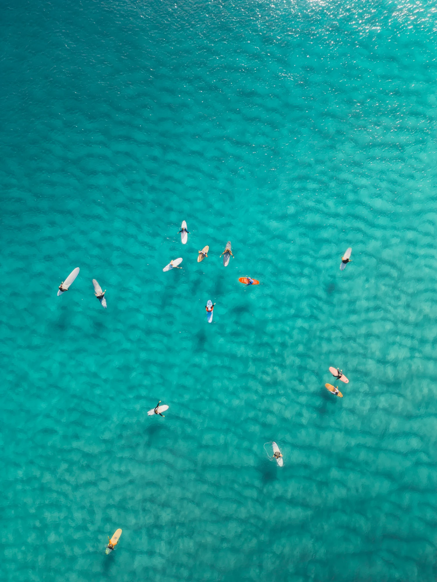Surfers and colourful boards are scattered above submerged rocks in clear turquoise water.