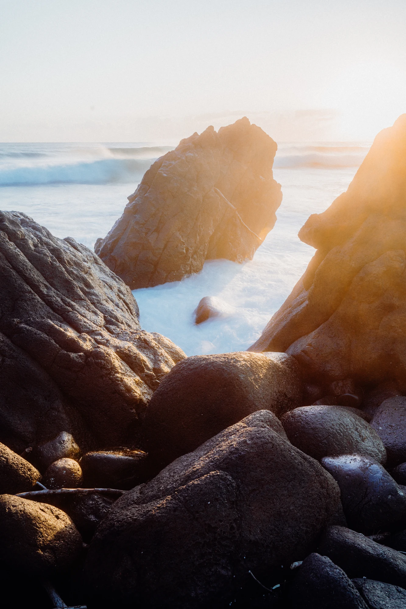 Golden sunlight shines between angular coastal rocks above rippled blue water.