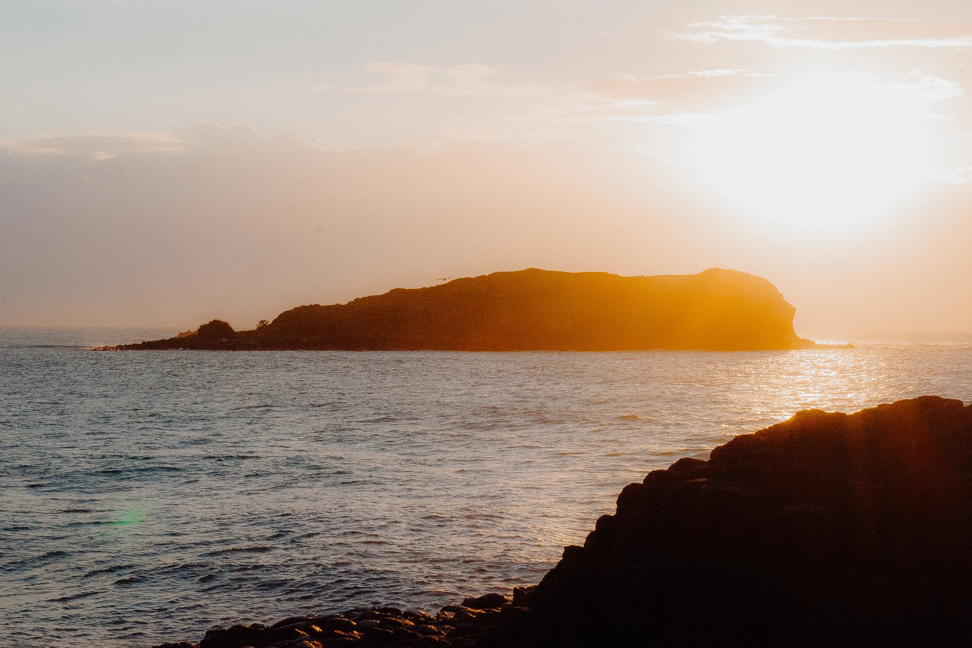 An offshore headland glows in orange backlight above the sea and dark foreground rock.