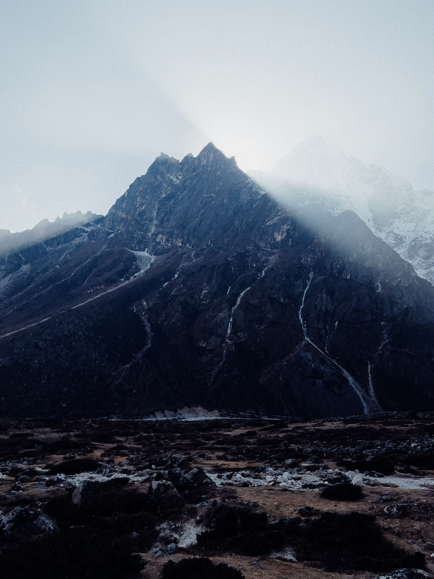 A dark jagged mountain emerges through drifting cloud above a rocky valley.