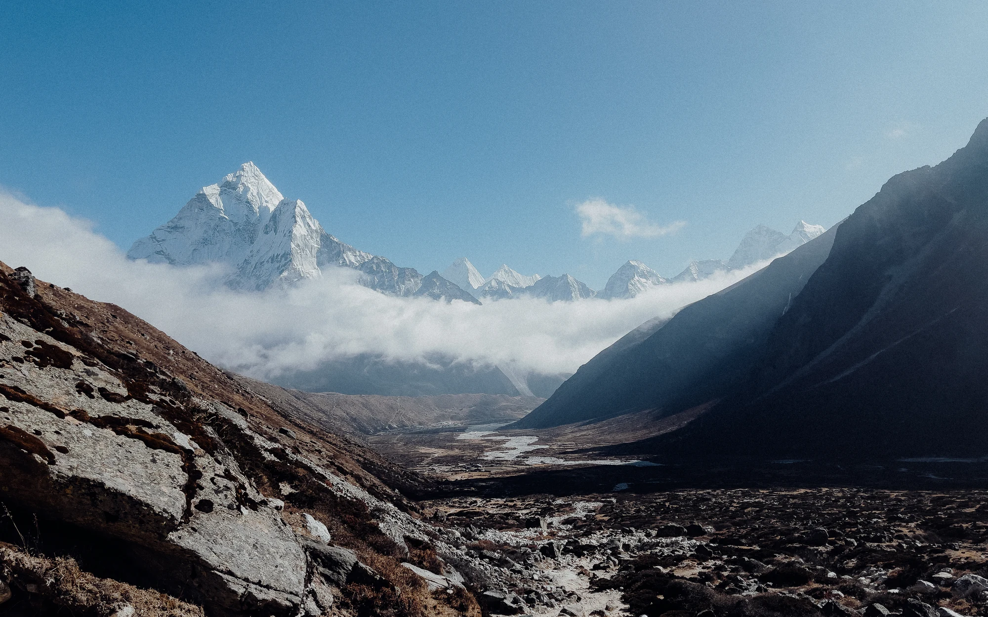 Snow peaks rise above a band of cloud beyond a rocky mountain valley.