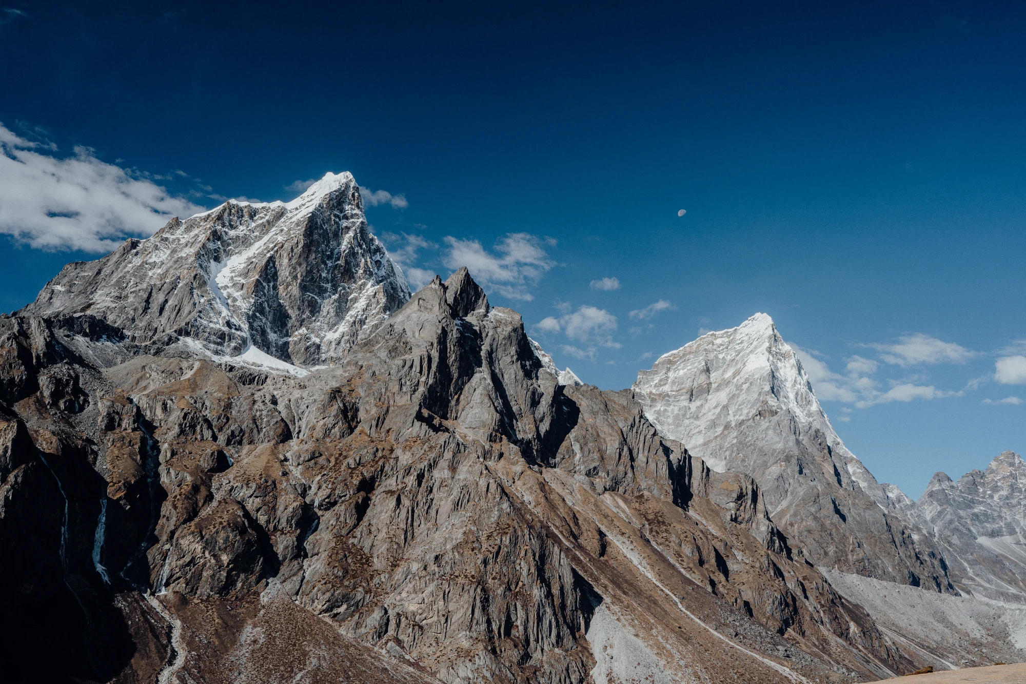 A pale moon hangs above angular snow-dusted mountain ridges beneath a deep blue sky.