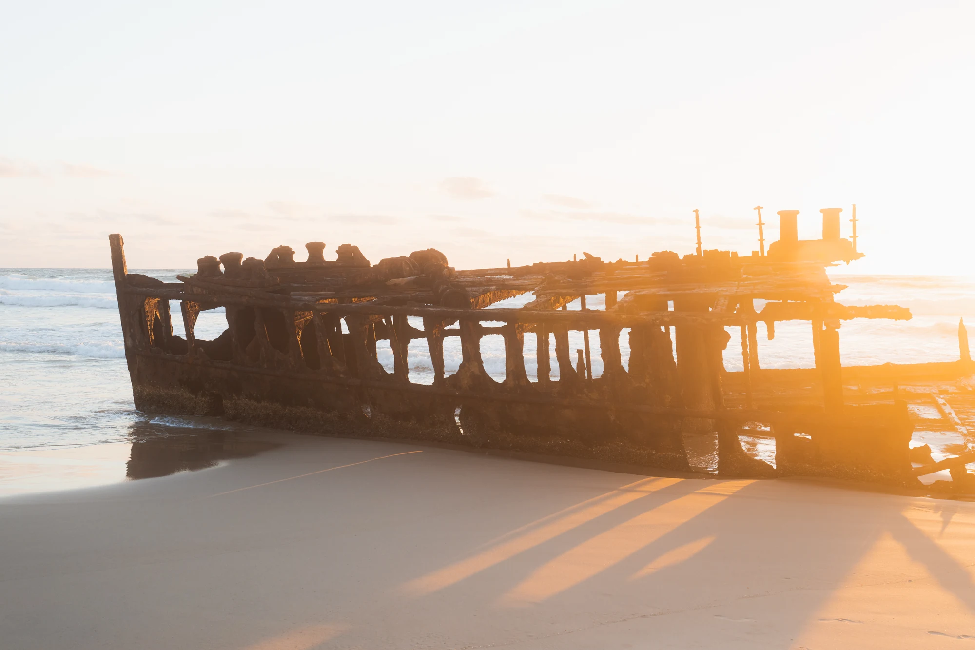 Golden sunlight shines through the rusted frame of a shipwreck on an empty beach.