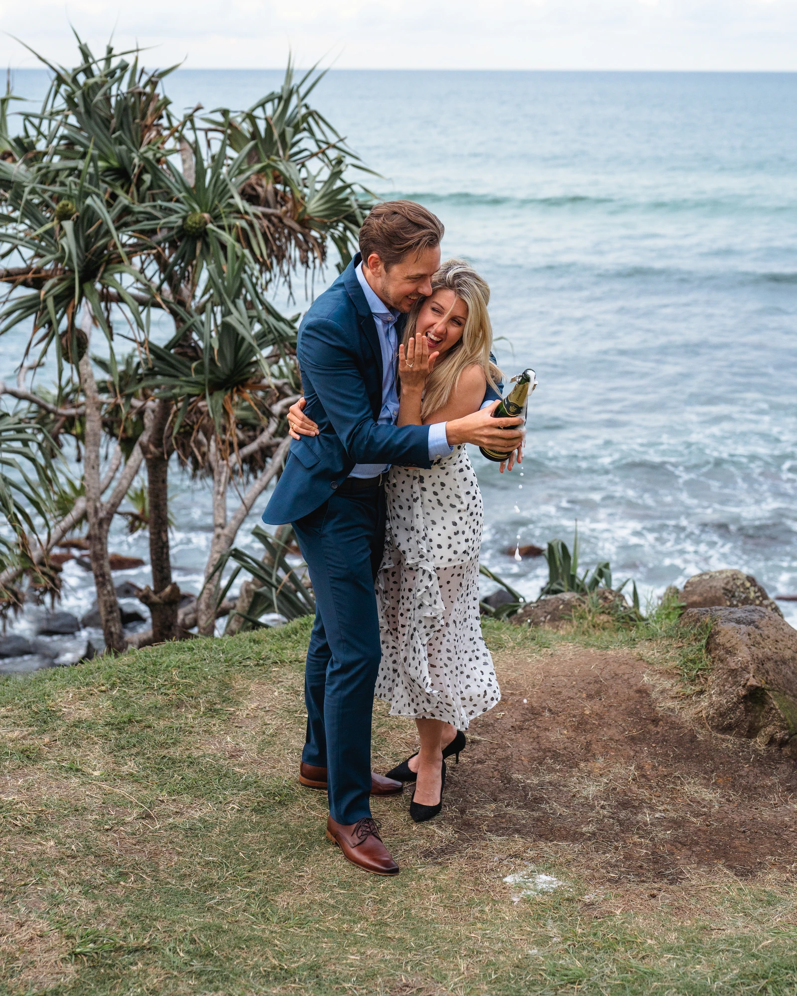 A couple hold each other and a champagne bottle beside the sea.