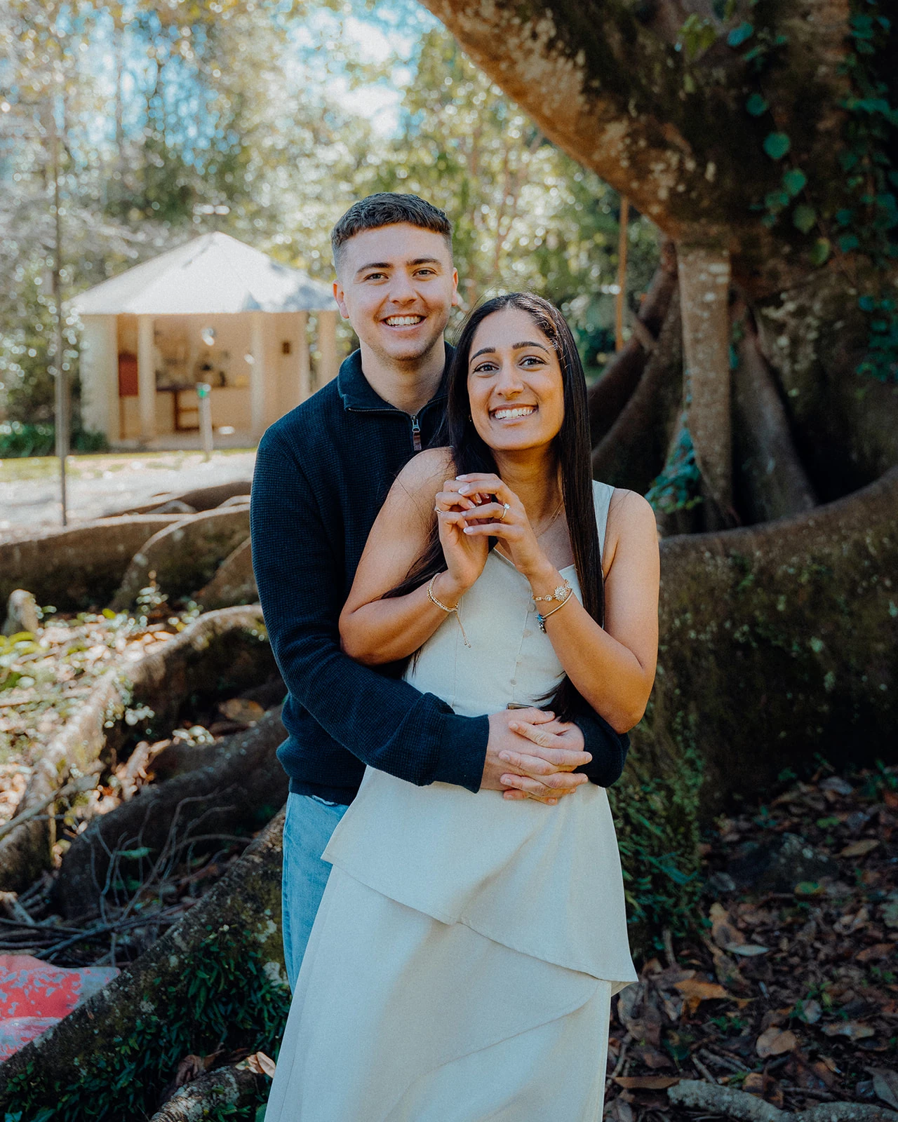 A smiling couple embrace beneath a tree.