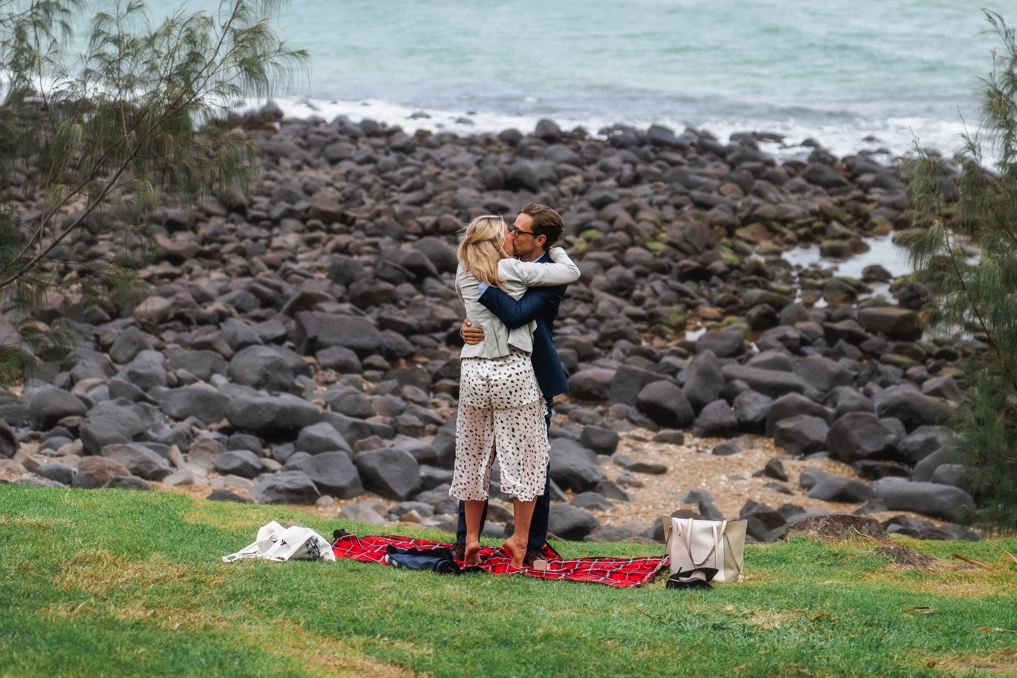 A couple embrace beside their picnic blanket after a coastal proposal.