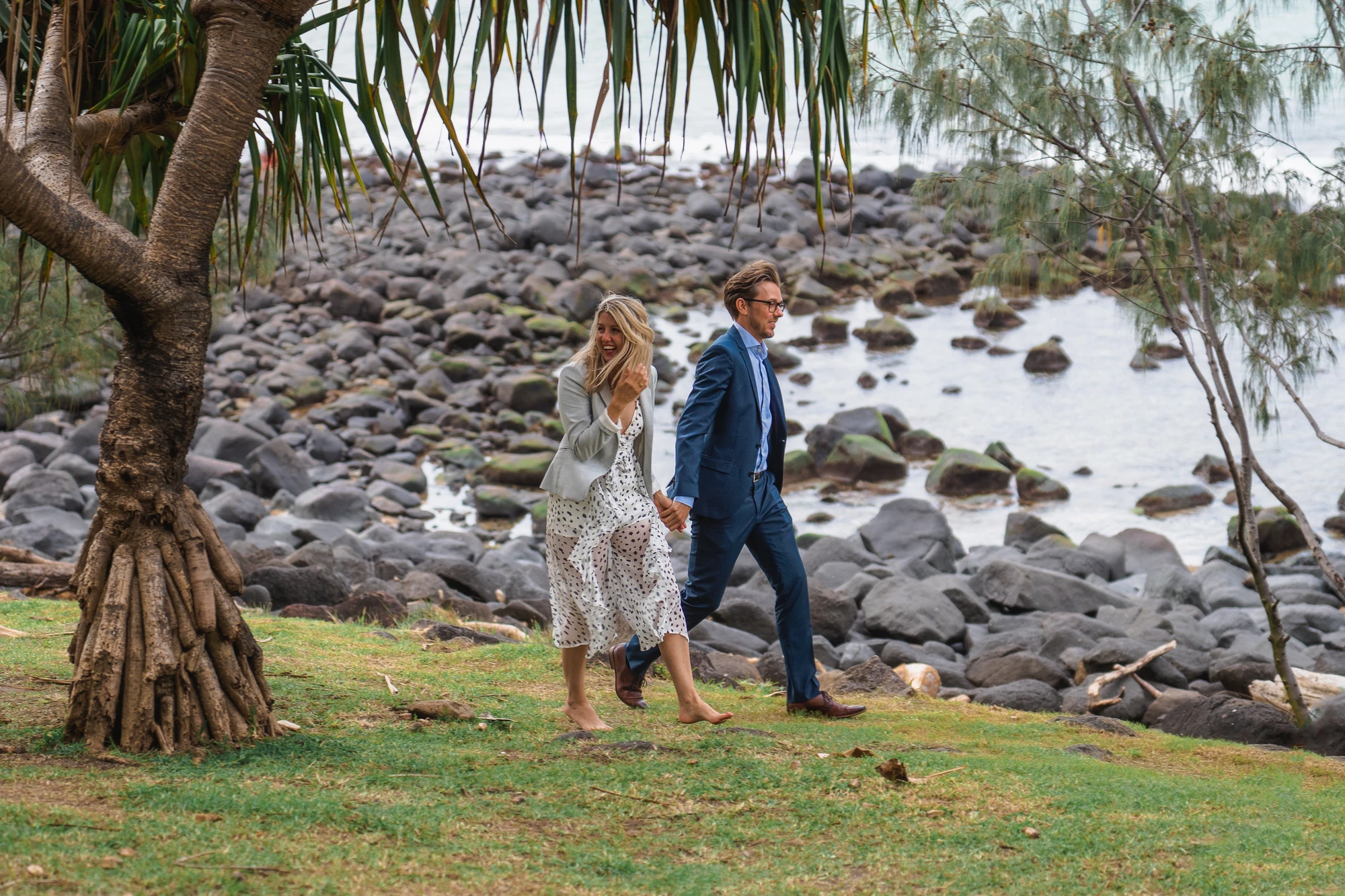 A couple walk hand in hand beneath pandanus trees beside the coast.
