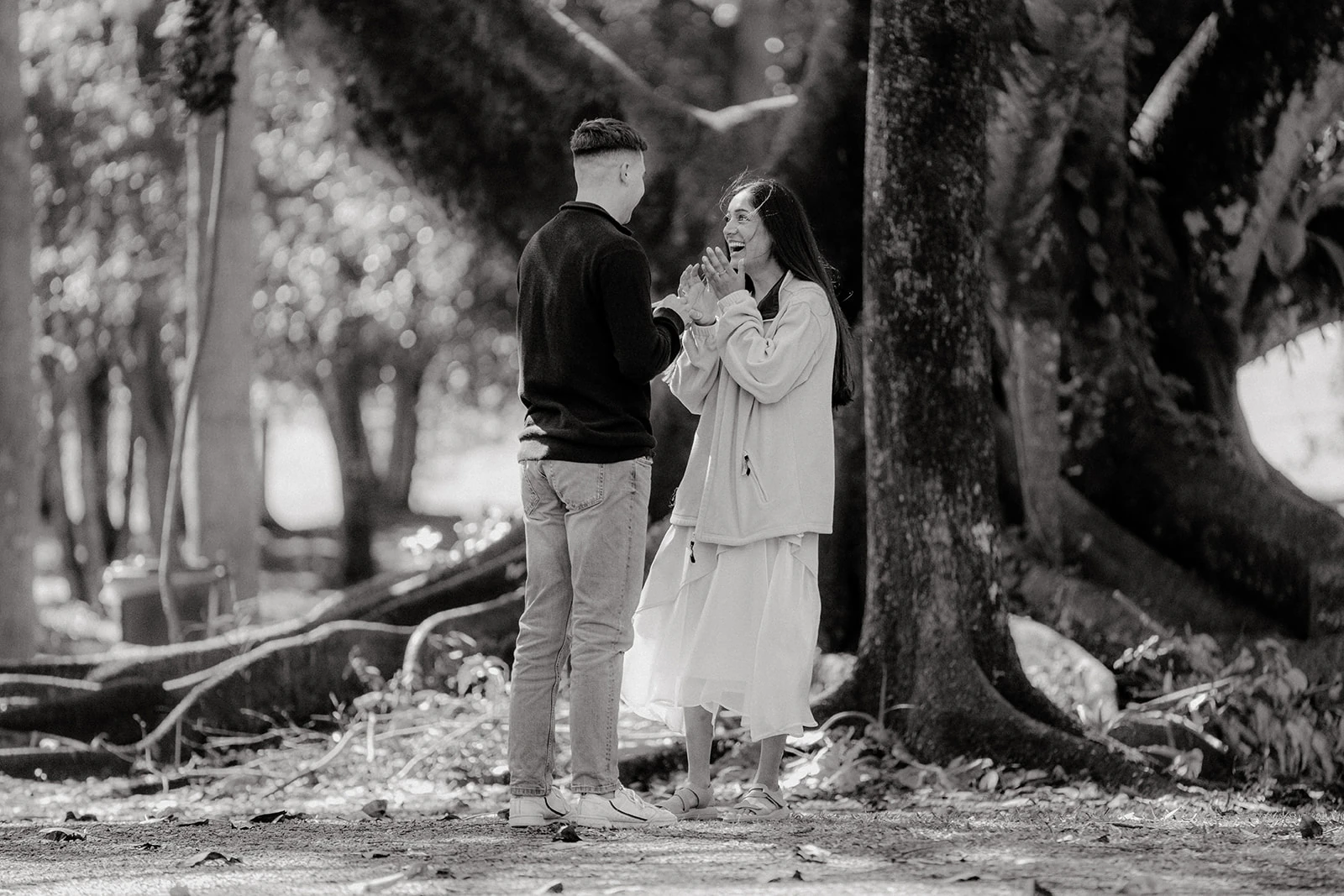 A couple laugh together beneath a large tree in black and white.