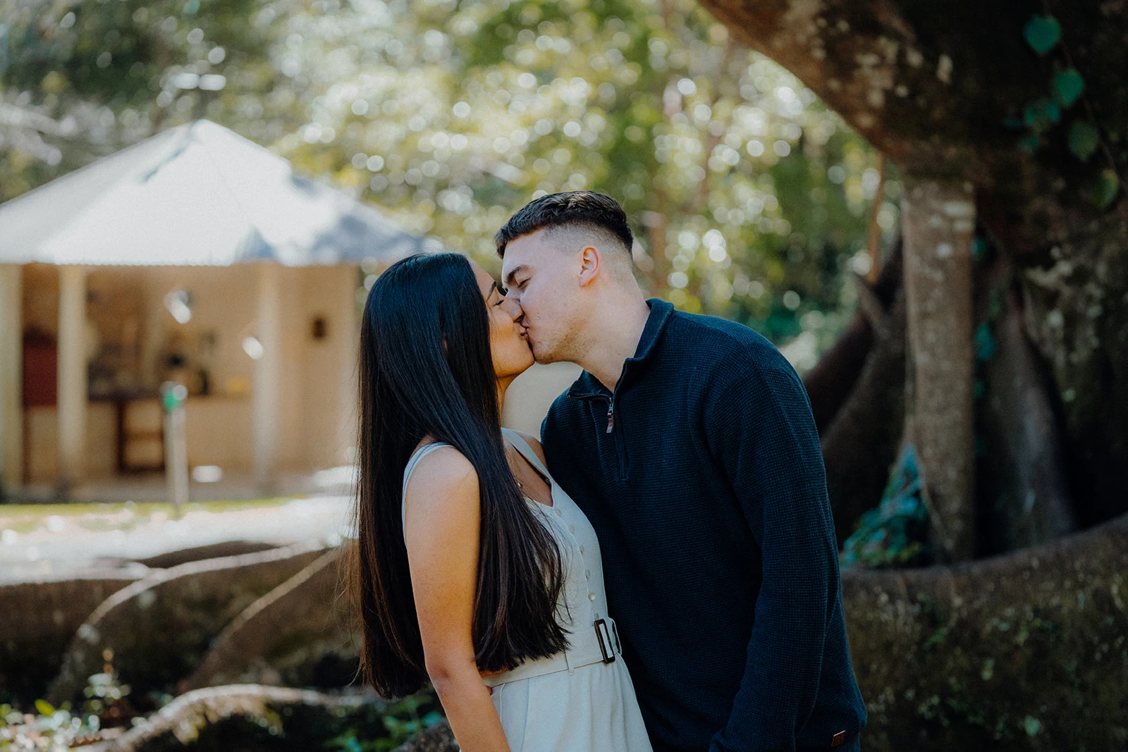 A couple kiss in the shade with a small chapel behind them.