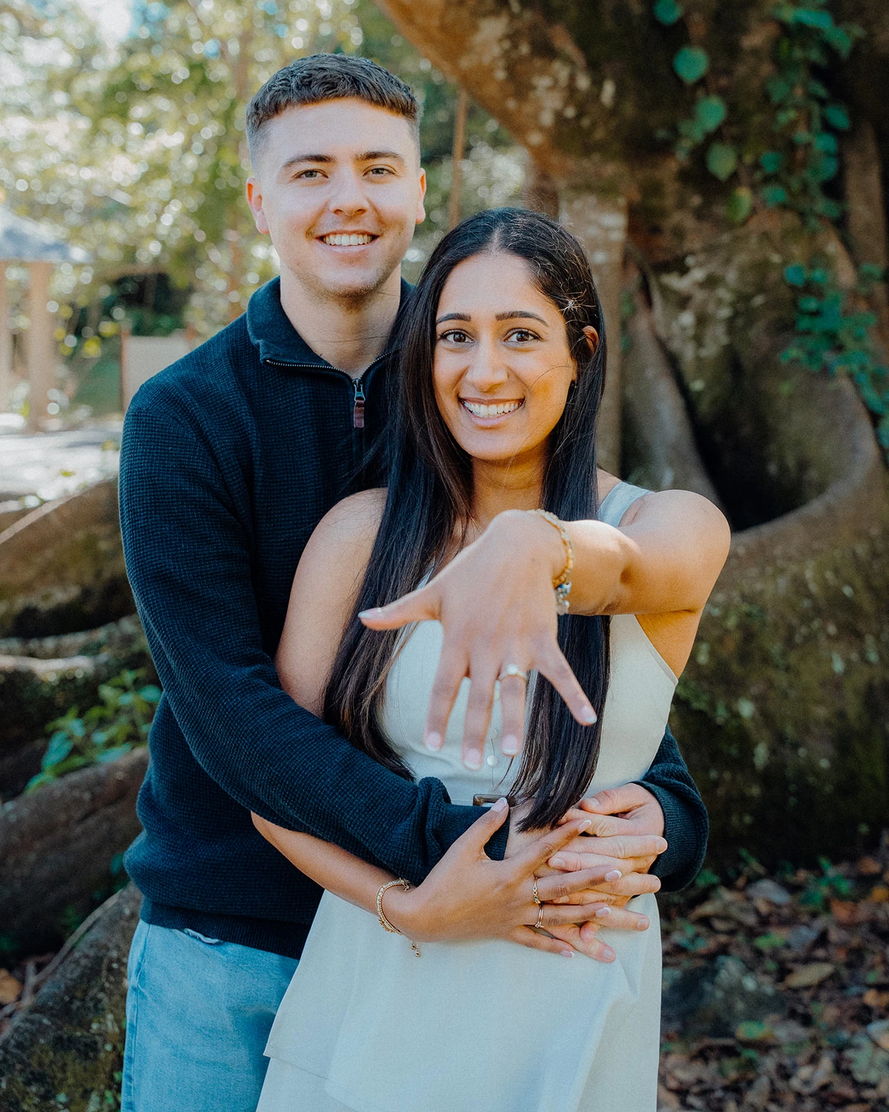 A woman holds out her ring hand while her partner embraces her from behind.