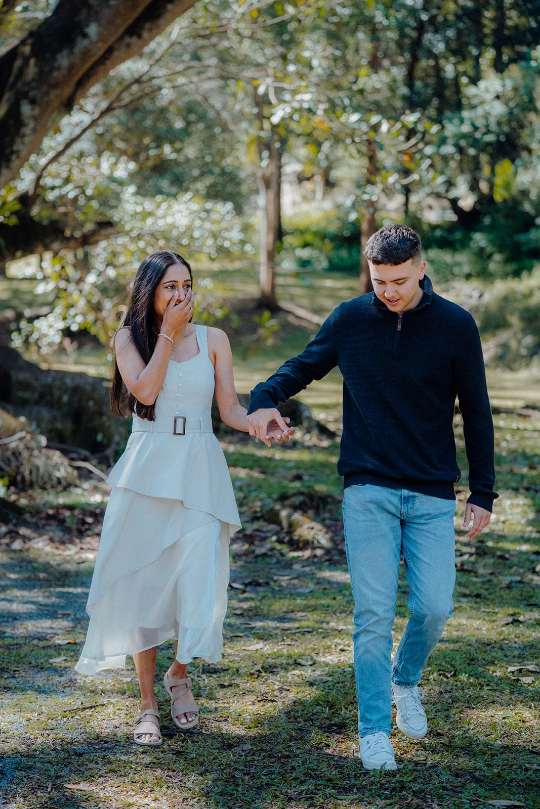 A couple walk toward the camera, holding hands beneath the trees.