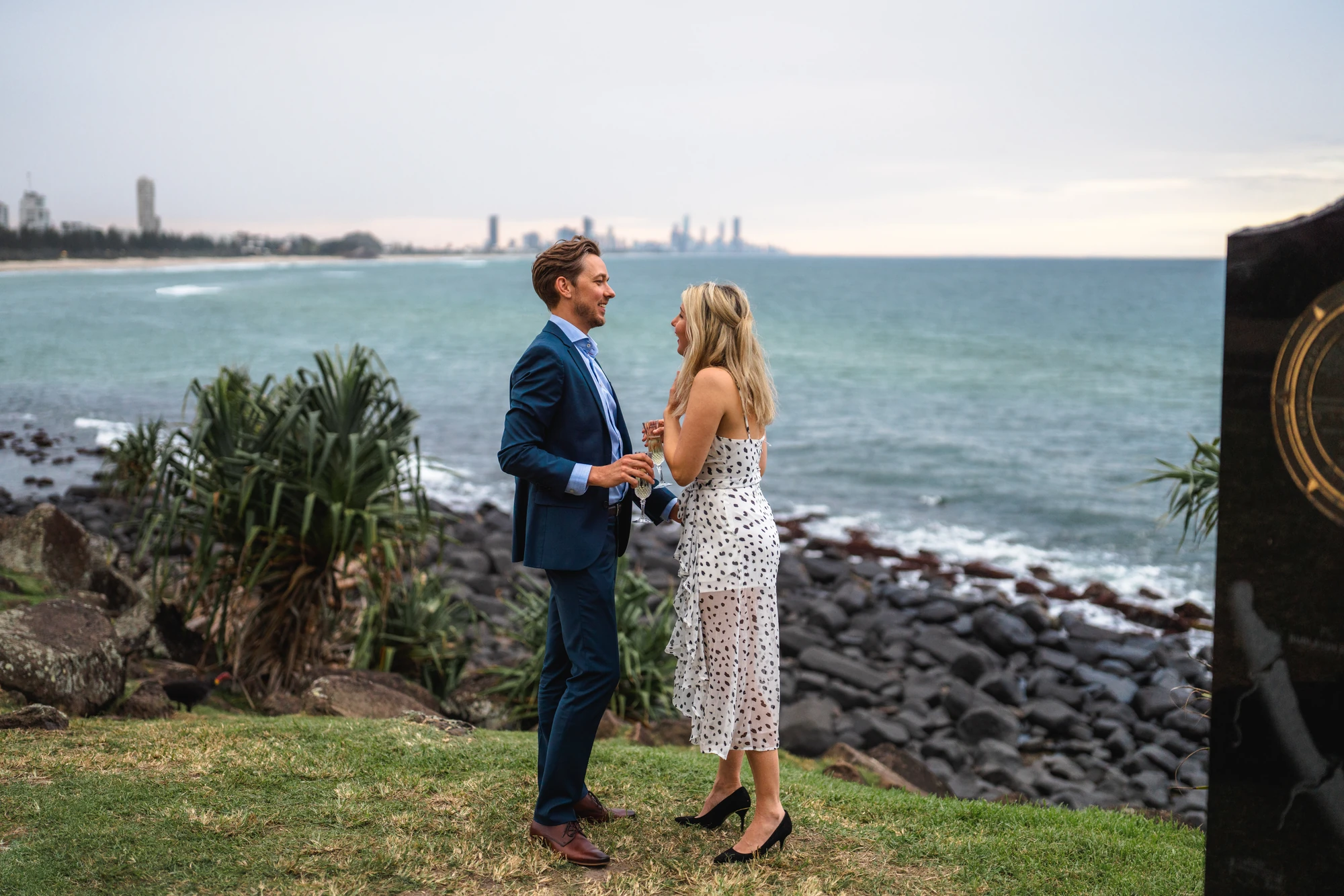 A couple face one another on a grassy headland with the city skyline across the bay.
