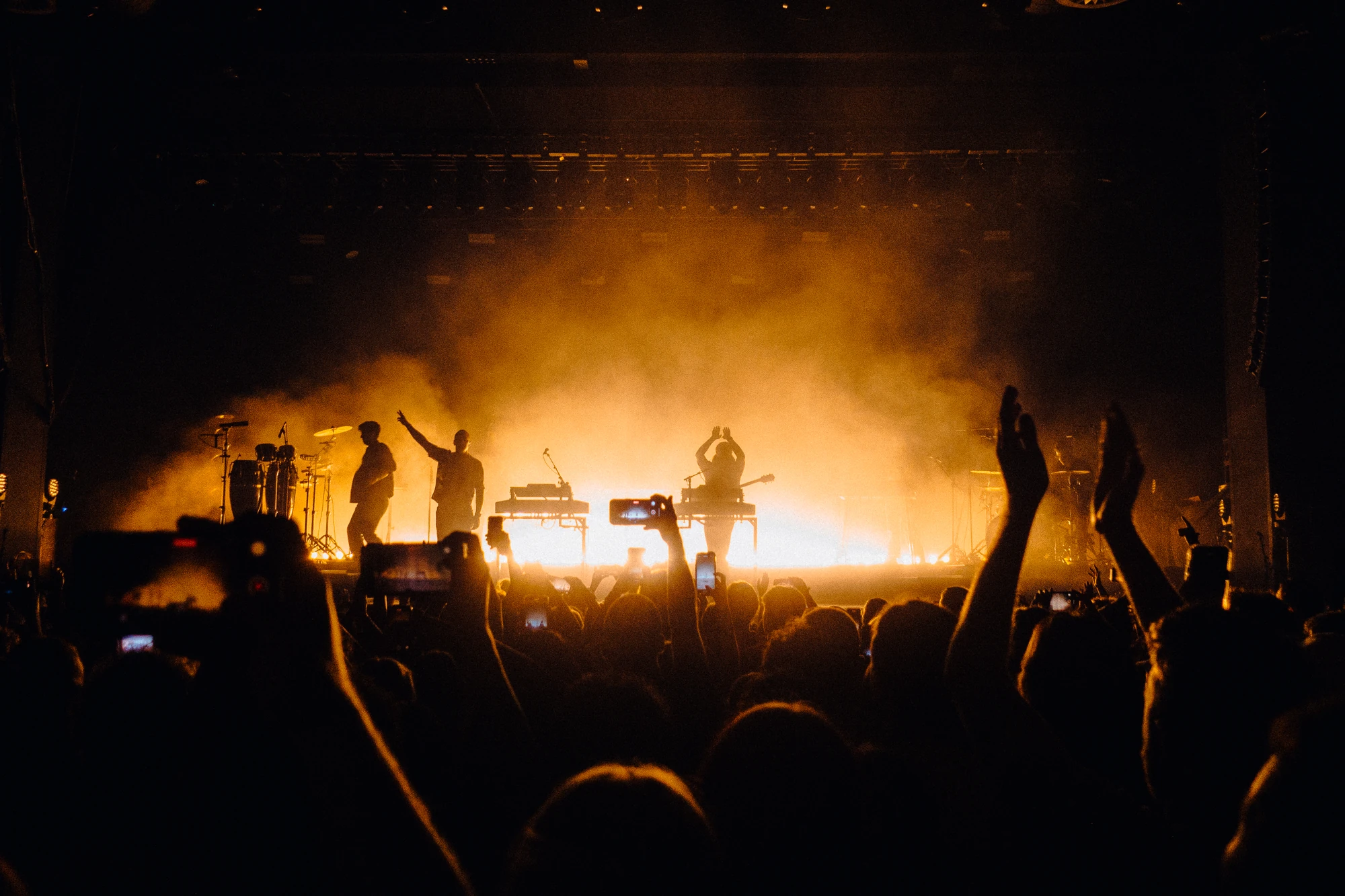 Musicians and raised audience hands are silhouetted against golden stage lights.