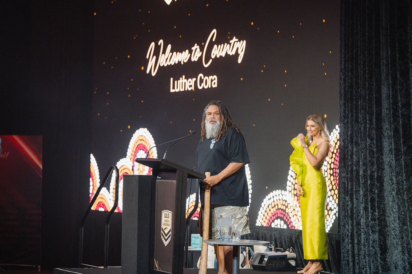 A speaker delivers a Welcome to Country beside an illuminated event screen.