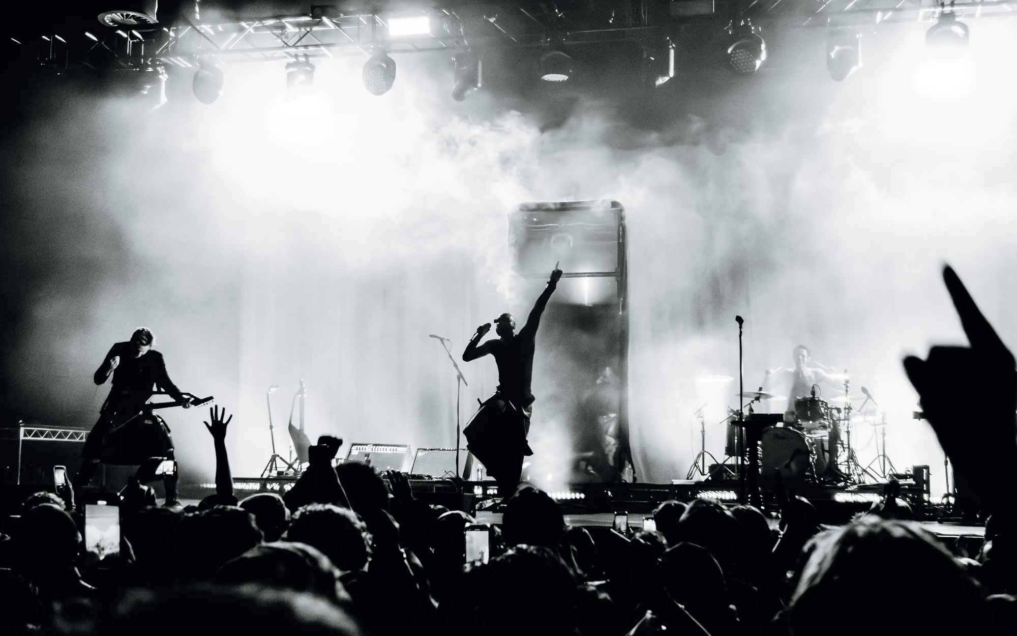 A singer raises an arm amid stage smoke and audience silhouettes in black and white.