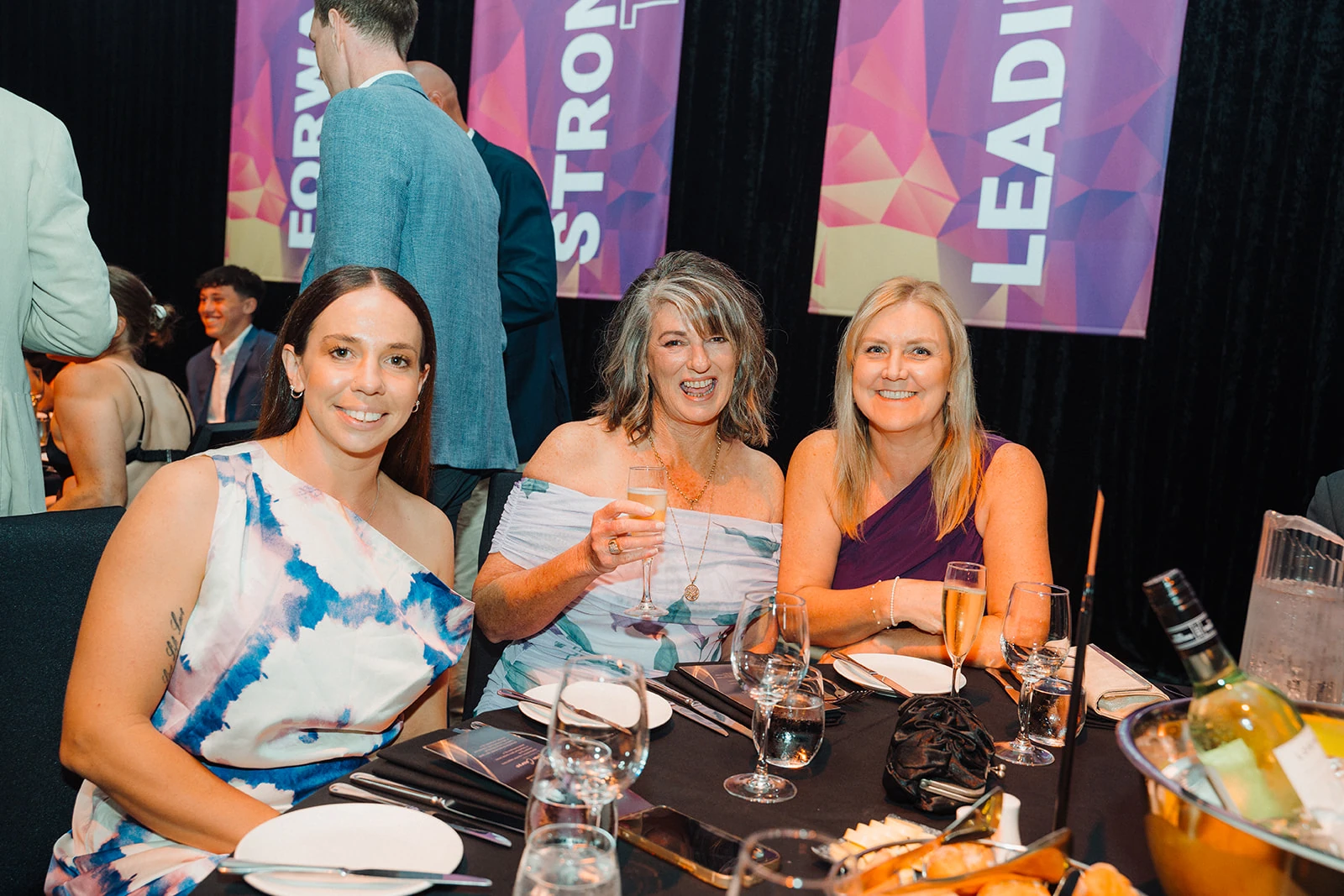 Three guests smile together at a table during an awards evening.