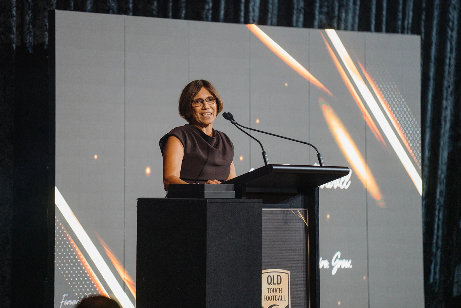 A woman speaks at a lectern against an illuminated gold-patterned screen.