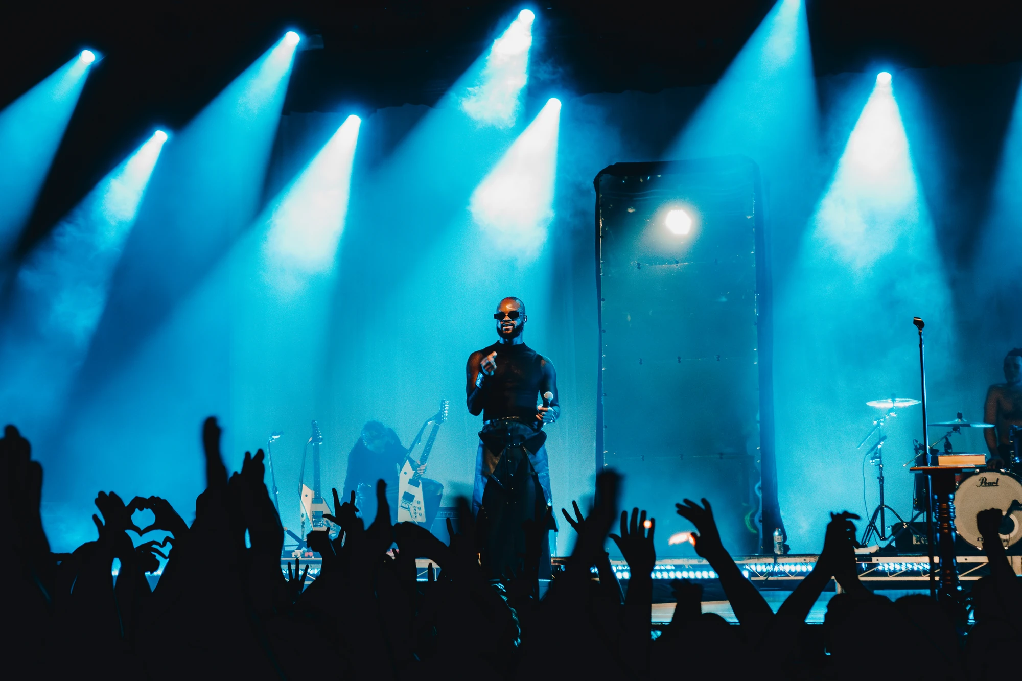 A singer faces a crowd of raised hands beneath cyan spotlights.