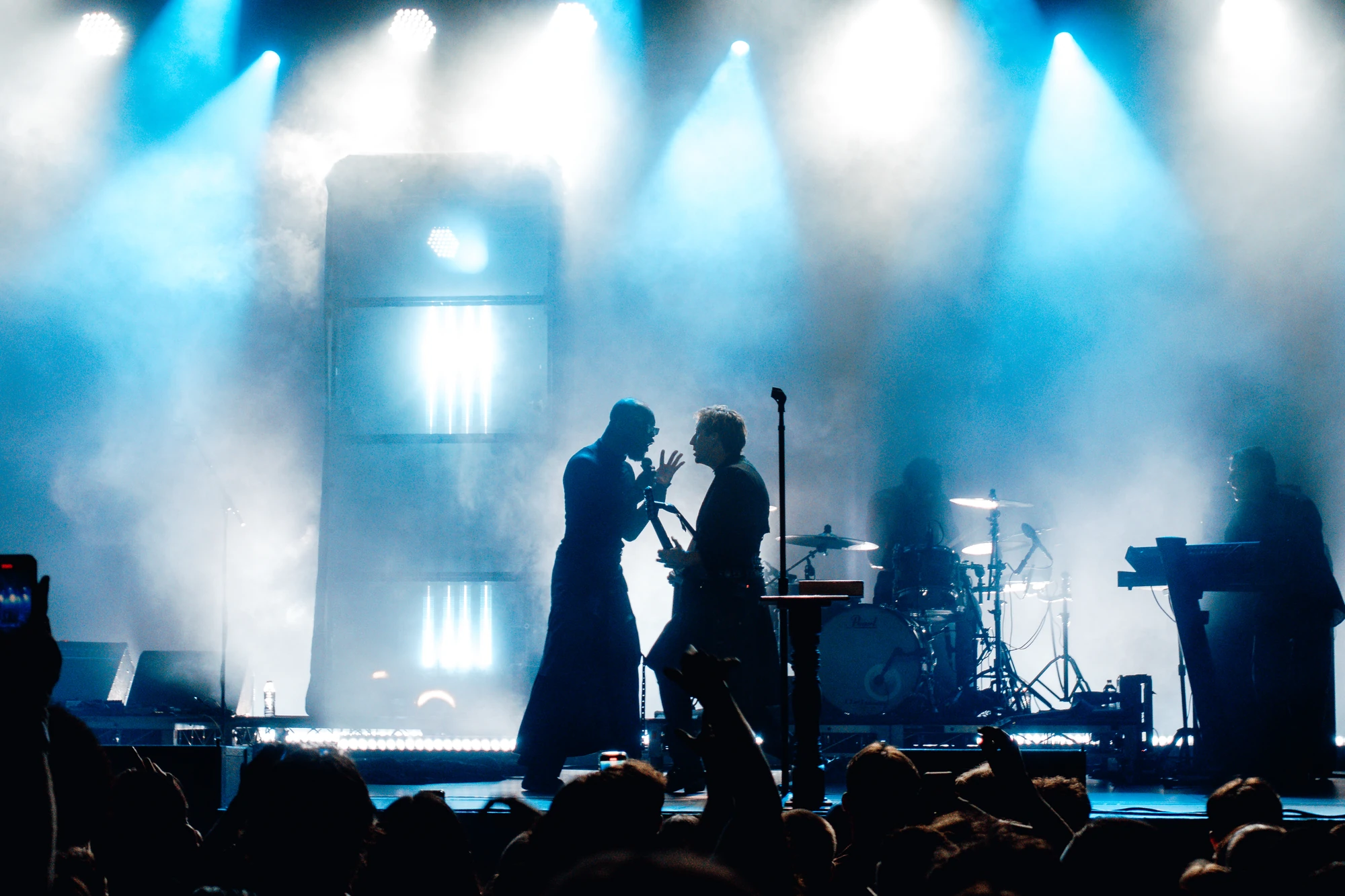 A singer and guitarist lean toward one another against white stage lights.
