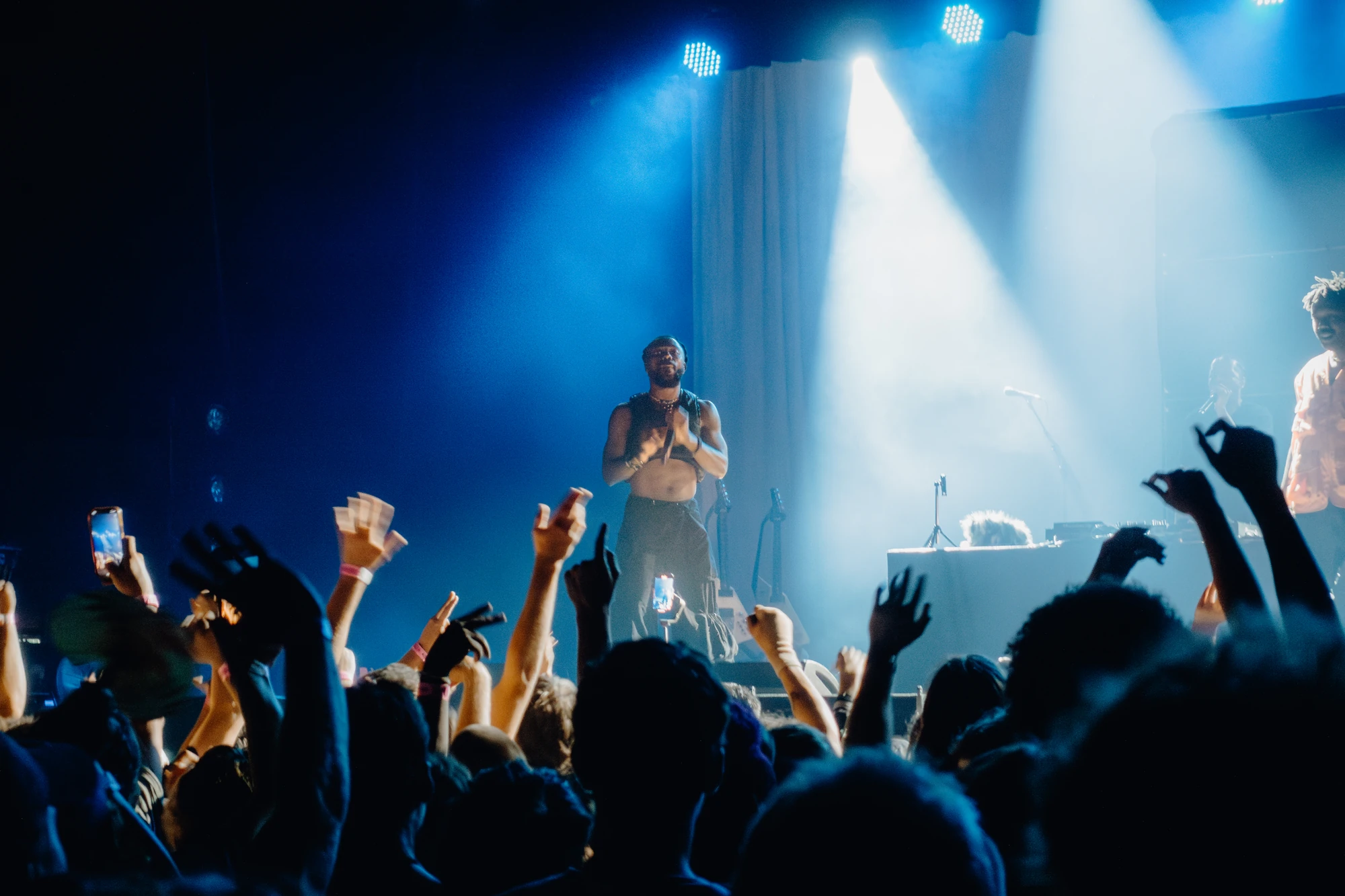 A performer faces an audience of raised hands beneath blue and white spotlights.