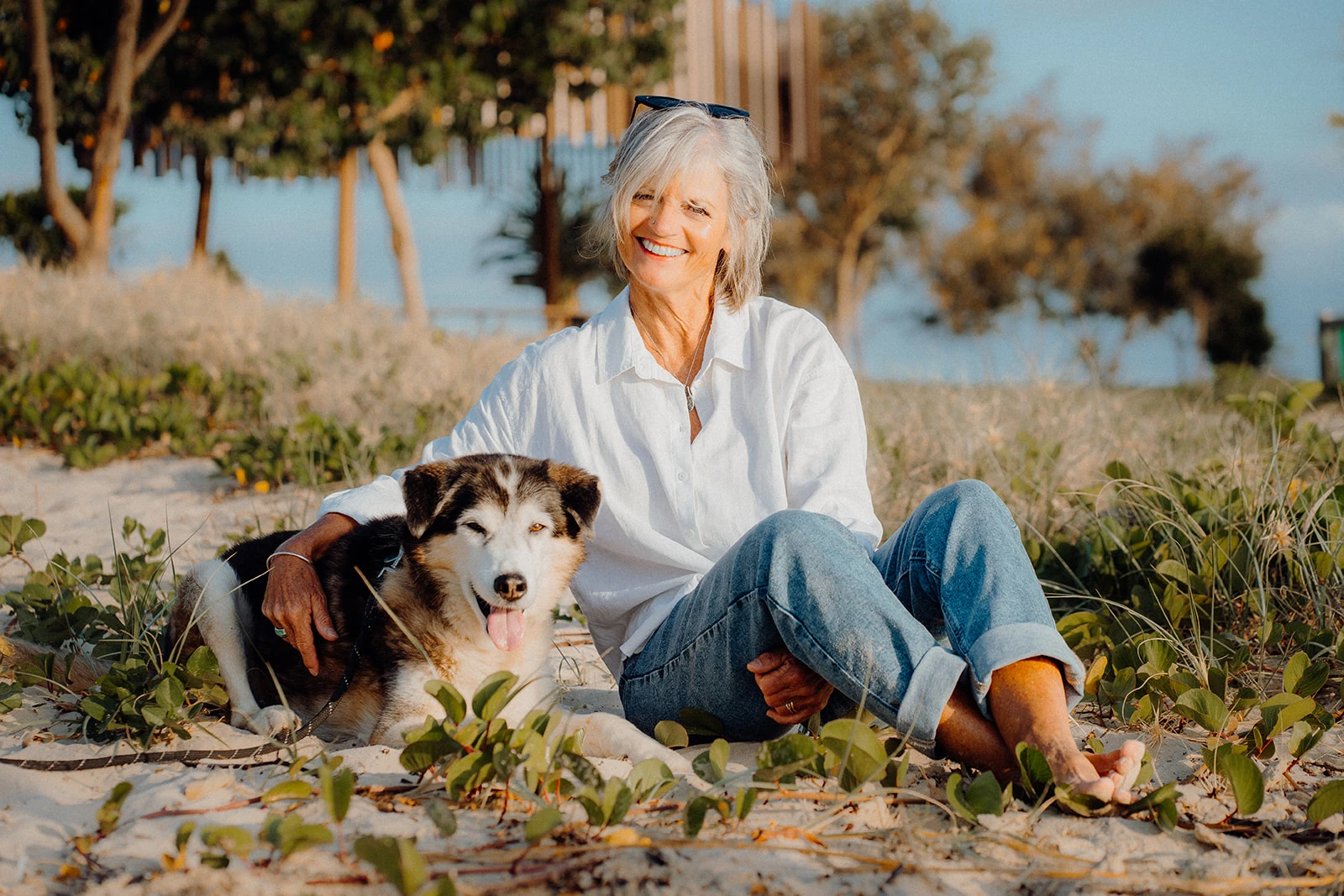 A woman sits smiling beside her dog among beach plants.