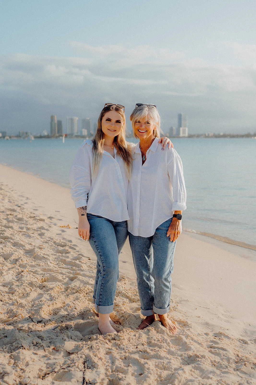 Two women stand arm in arm beside the water.
