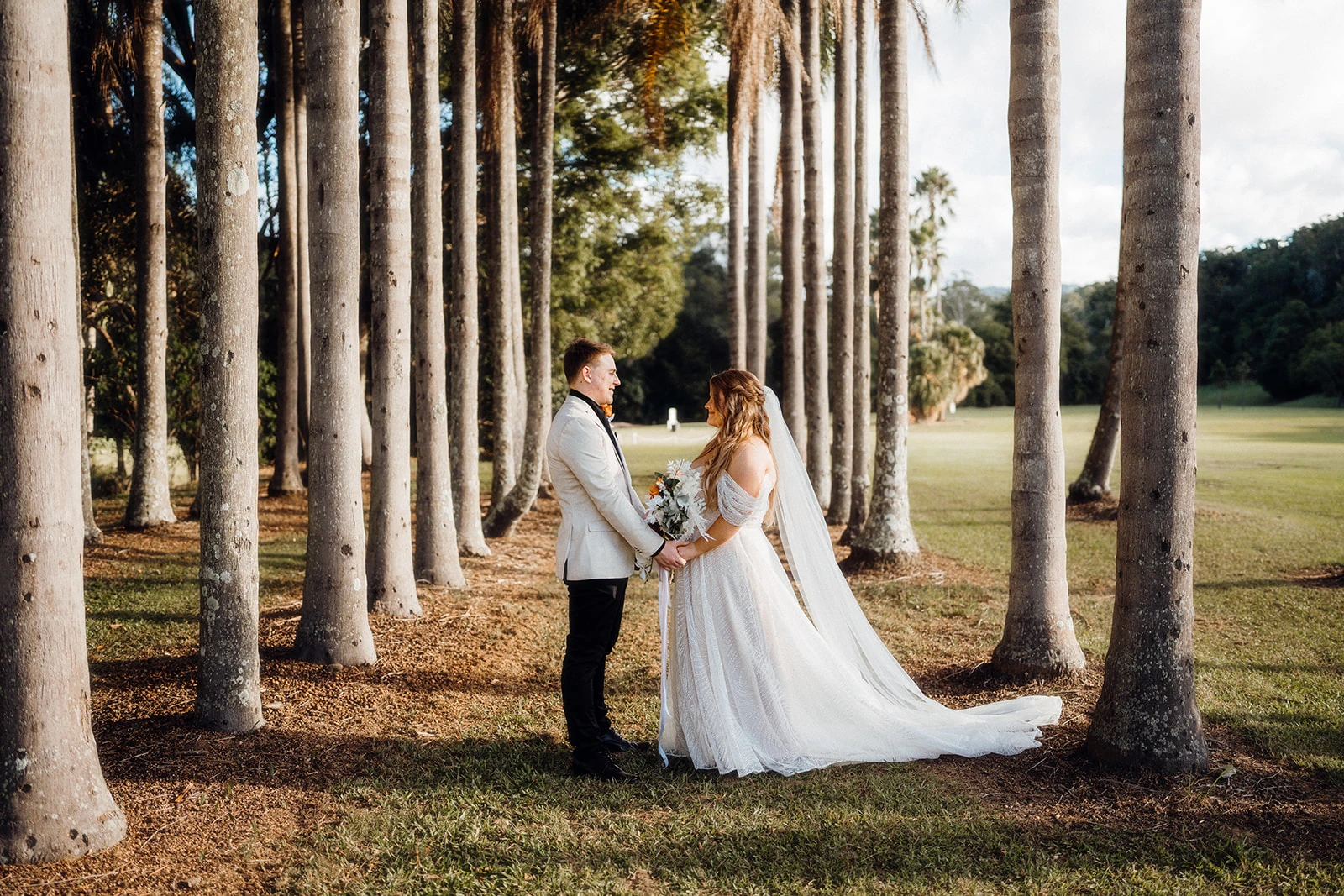A wedding couple face one another between tall palm trunks.