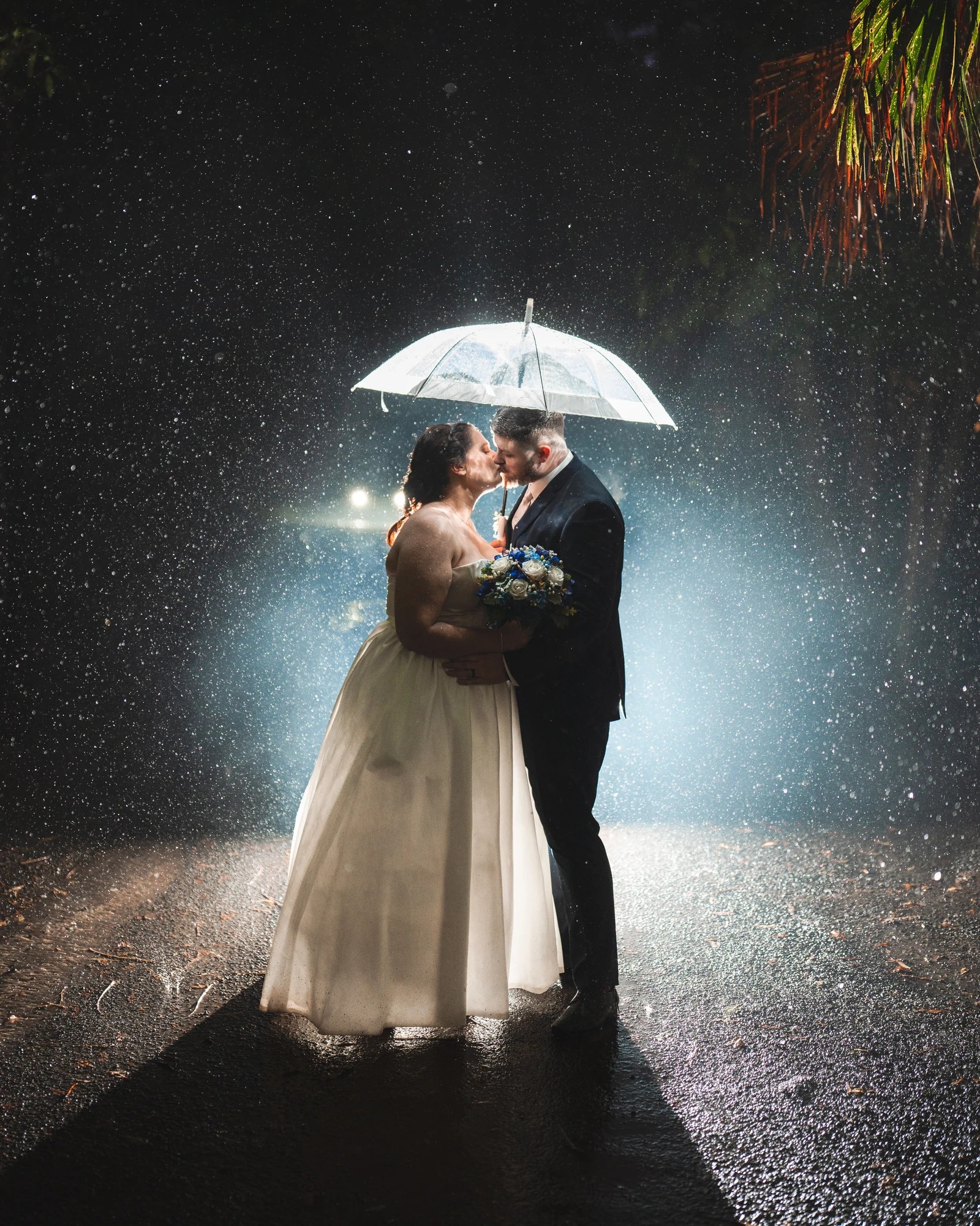 A wedding couple kiss beneath a clear umbrella in the backlit rain.