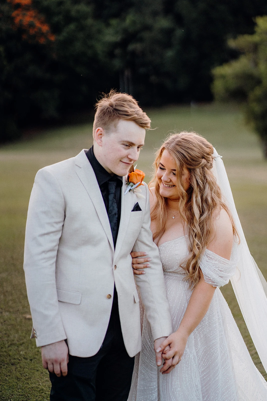 A bride and groom smile at one another while holding hands on a lawn.