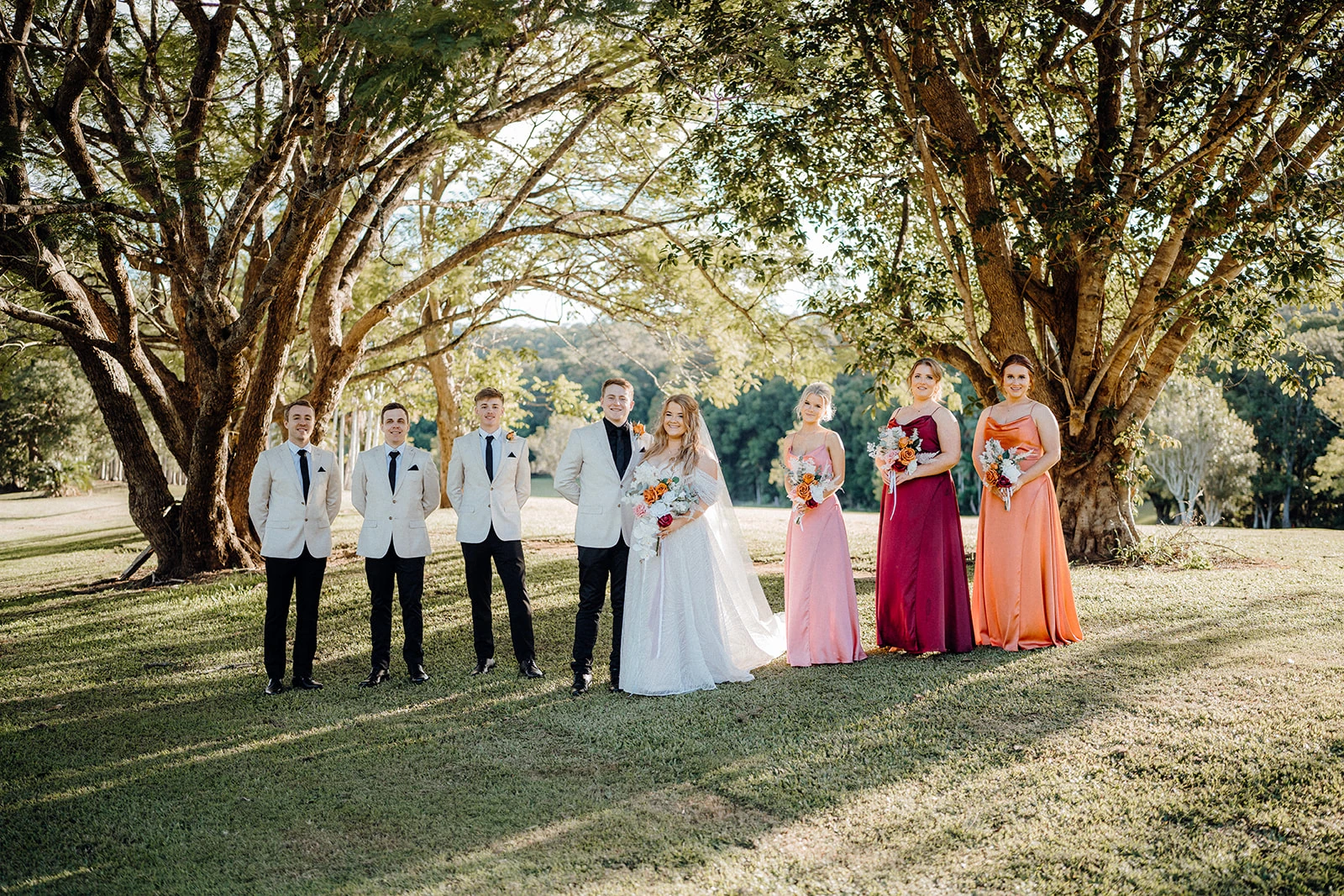 A wedding party stands beneath an avenue of trees.