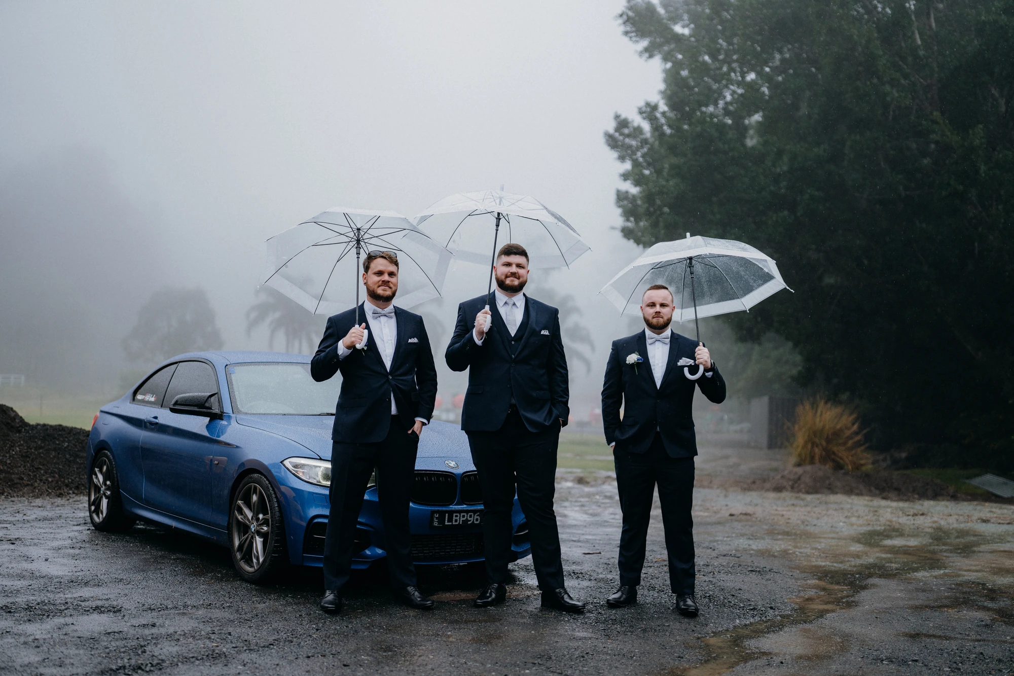 Three men in suits hold clear umbrellas beside a car in the mist.