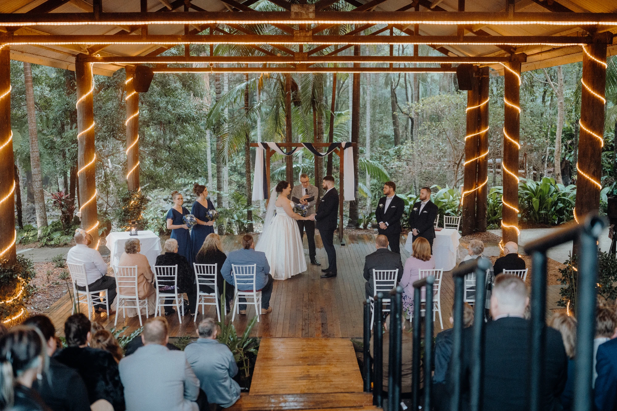 Guests watch a ceremony beneath a timber roof and strings of lights.
